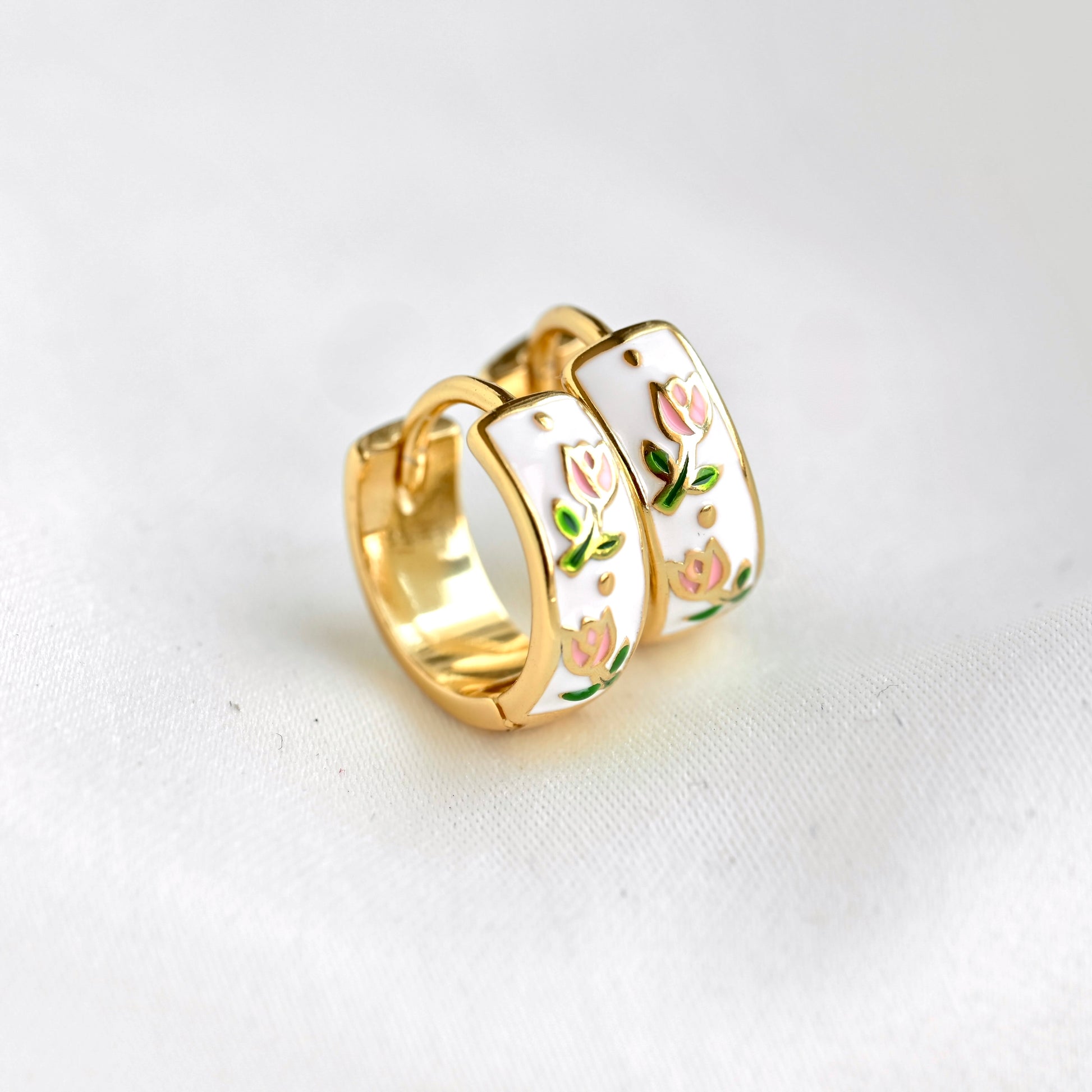 A pair of gold hoop earrings with white enamel panels decorated with pink tulips and green leaves, resting on a white textured fabric background.