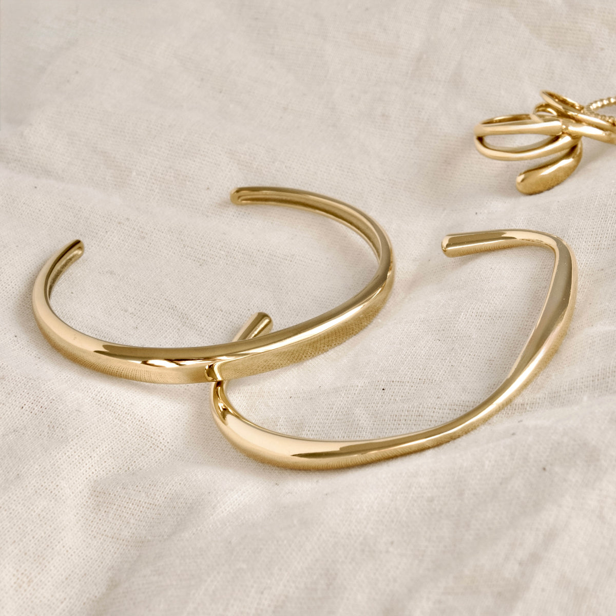 Gold-toned open cuff bracelets with a polished finish, displayed on a textured off-white fabric background.