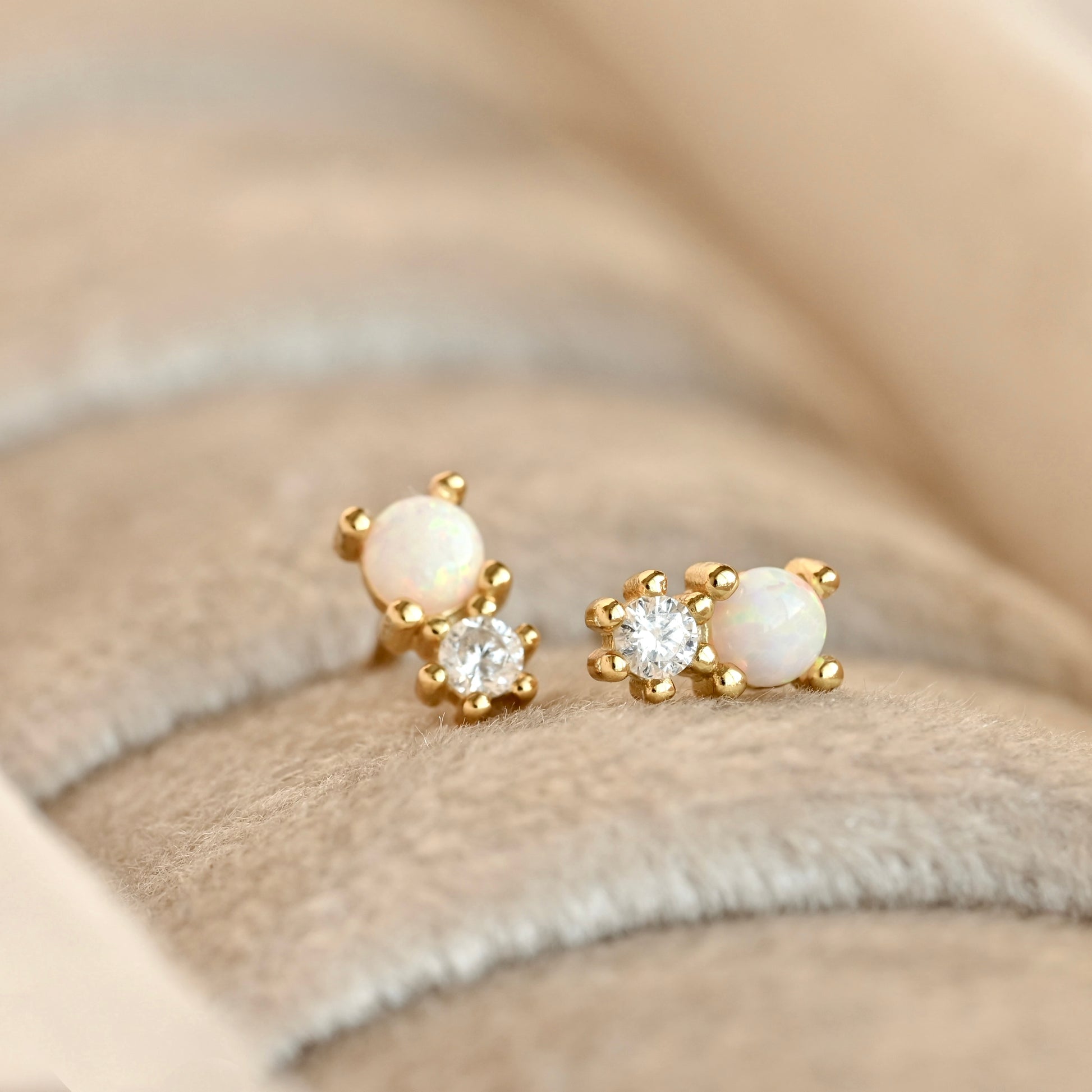 A pair of gold stud earrings with one round white opal and one clear crystal per earring, displayed on a beige textured velvet jewelry box.