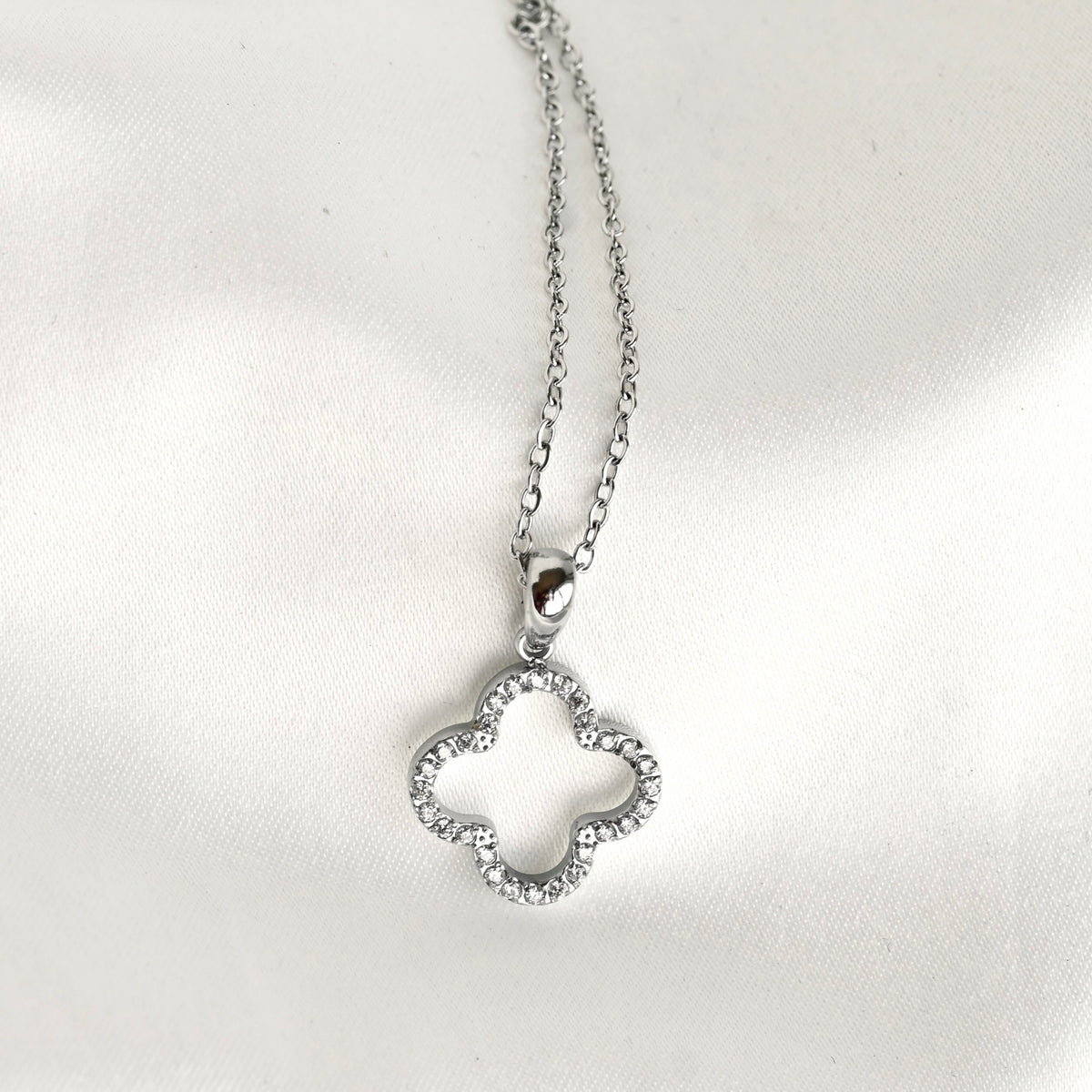 A silver-toned necklace with a four-leaf clover pendant, photographed on a white satin fabric background.