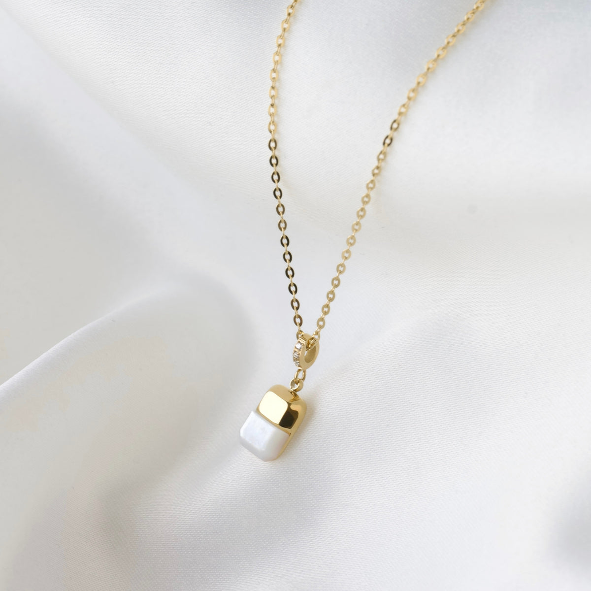 A delicate gold pearl necklace with a small capsule-shaped pendant, featuring a white pearl inlay on the bottom half and a polished gold top half, displayed on a soft white fabric background.