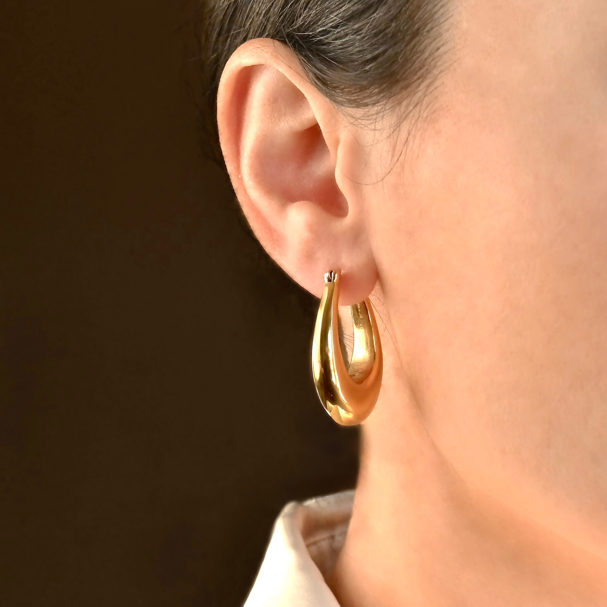 Chunky large gold teardrop hoop earrings worn by a person against a dark background