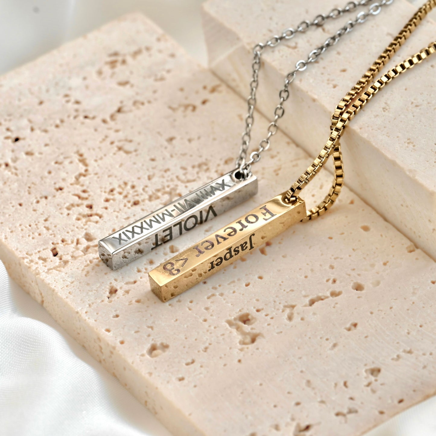 Two thick bar necklaces with engraved names on a textured surface