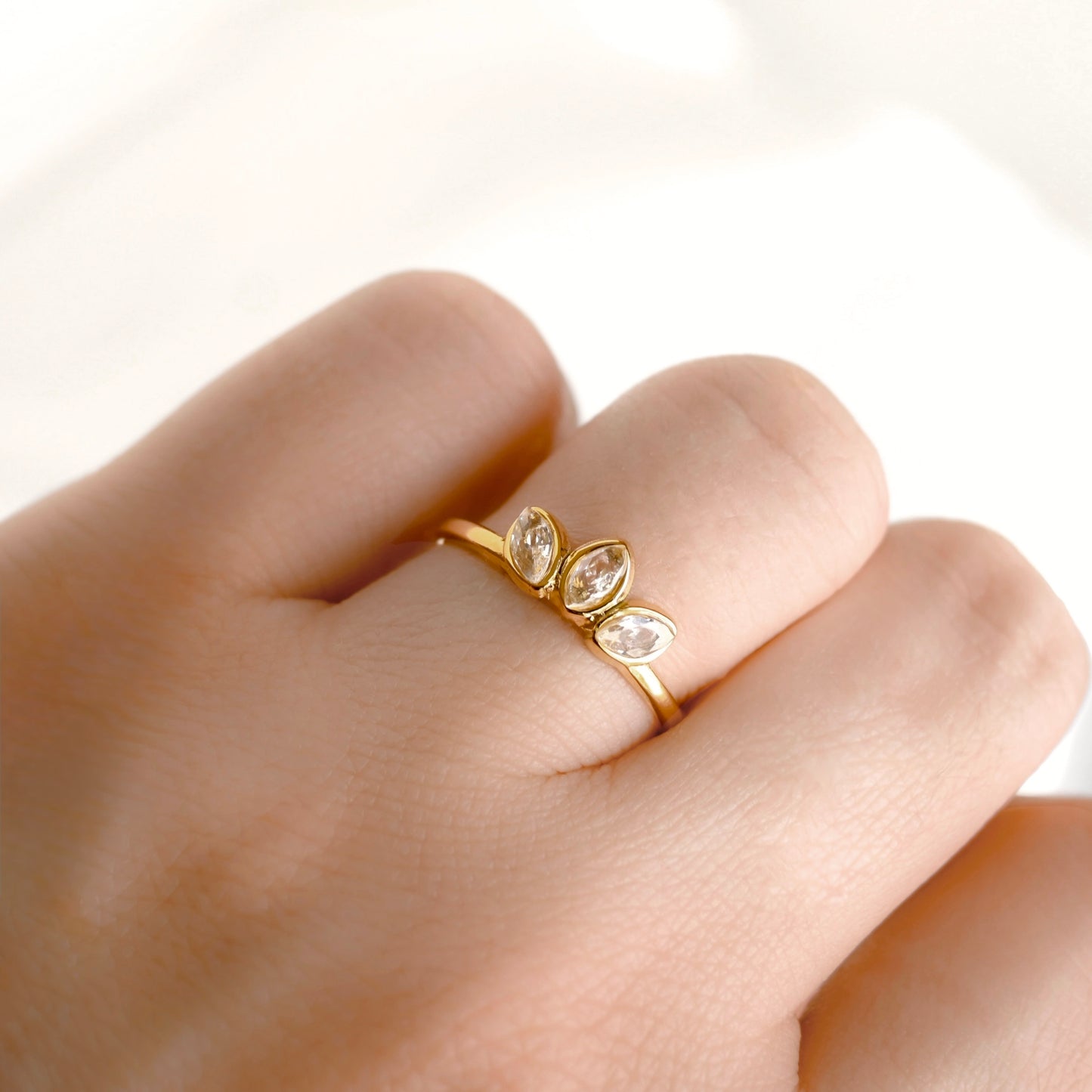 A close-up of a hand wearing a delicate gold lotus ring with three marquise-cut clear stones set in a row.