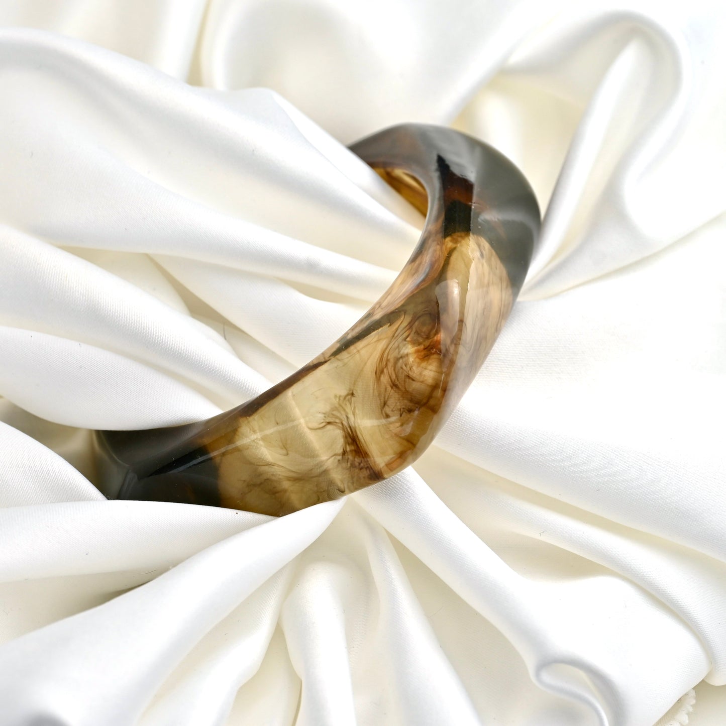 A chunky curved resin bangle with a marbled brown and black pattern, displayed on draped white satin fabric.