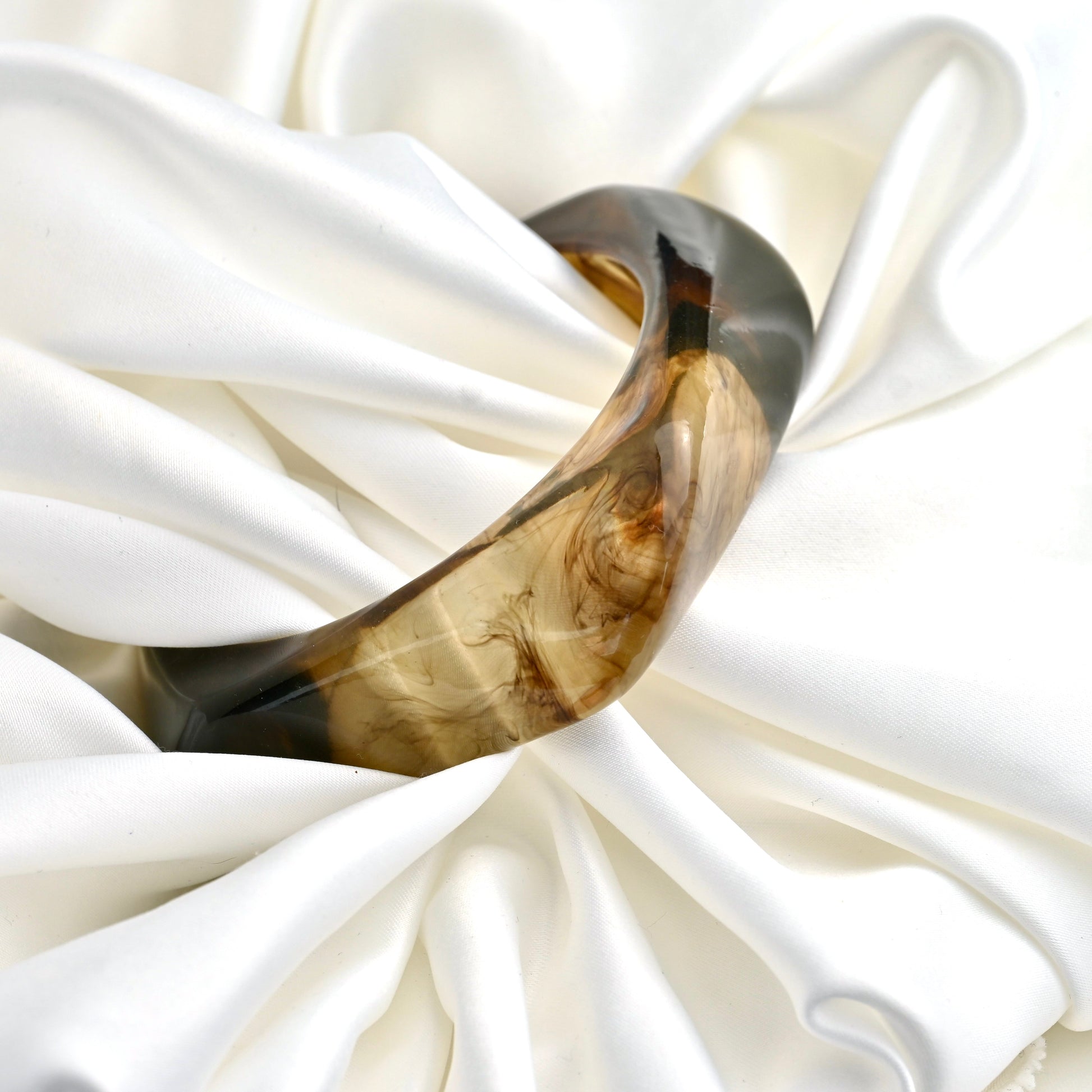 A chunky curved resin bangle with a marbled brown and black pattern, displayed on draped white satin fabric.