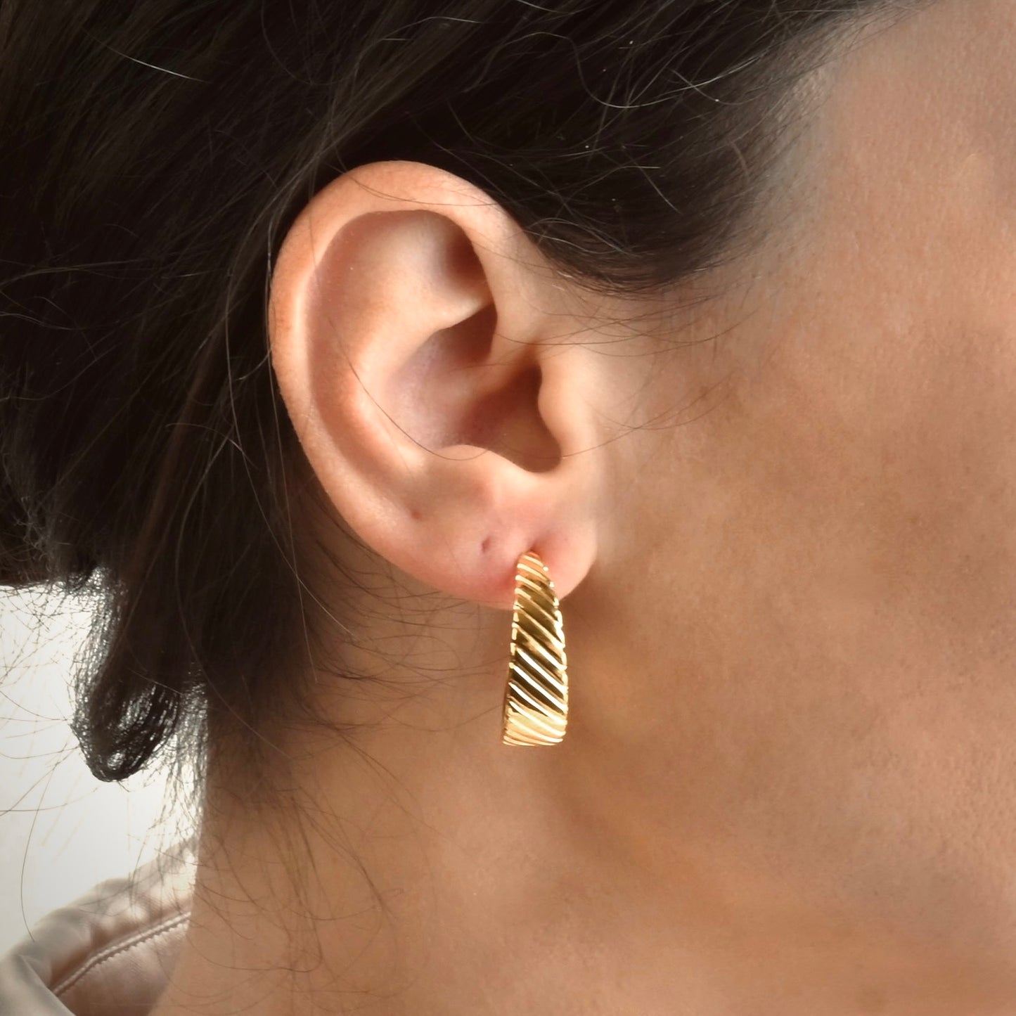 Tapered gold hoop earring  with ribbed texture worn by a person with dark hair.