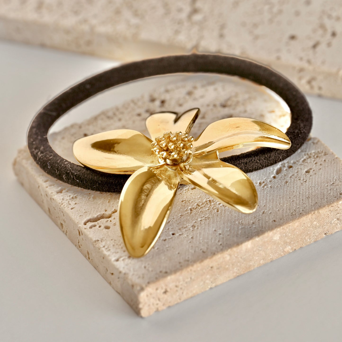 A black hair tie with a hibiscus flower accent  resting on a dark brown elastic band, displayed on a light beige stone block.