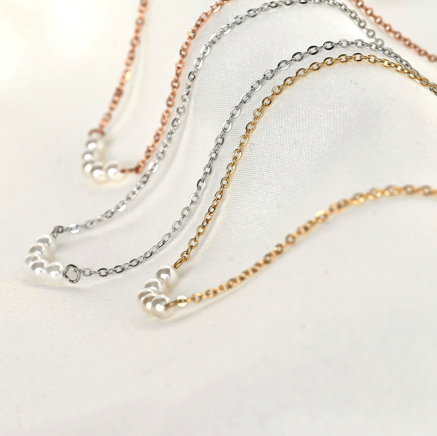 Side close-up view of three pearl necklaces in gold, silver, and rose gold on a light background