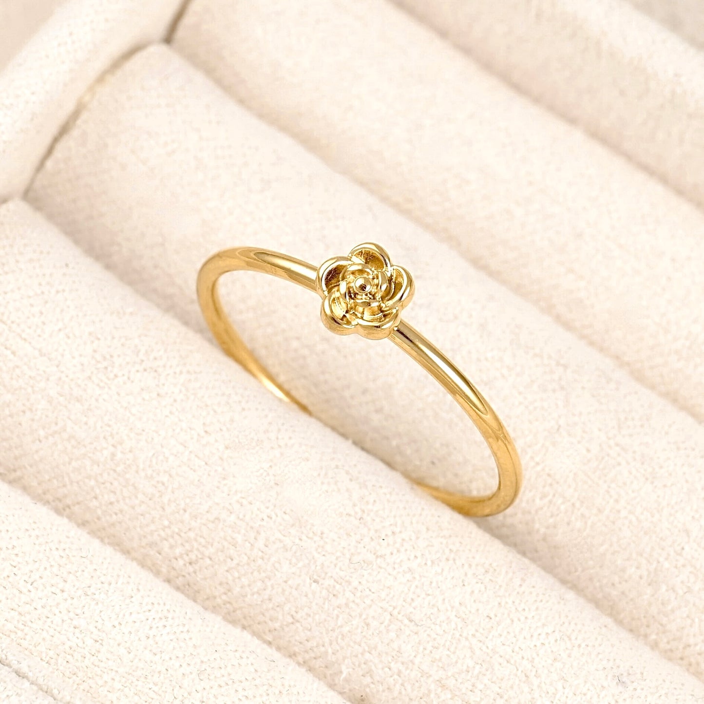 A delicate yellow gold ring featuring a small, textured flower motif at the center, displayed on a cream-colored textured ring cushion.