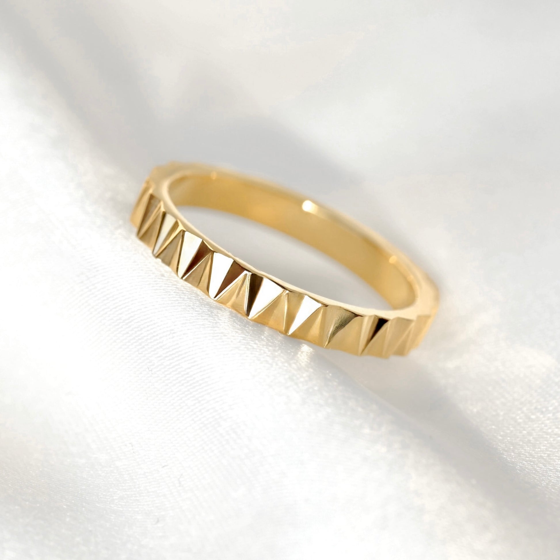 A gold stacking ring with a textured, faceted triangular pyramid pattern around the band, resting on a soft white satin fabric background.