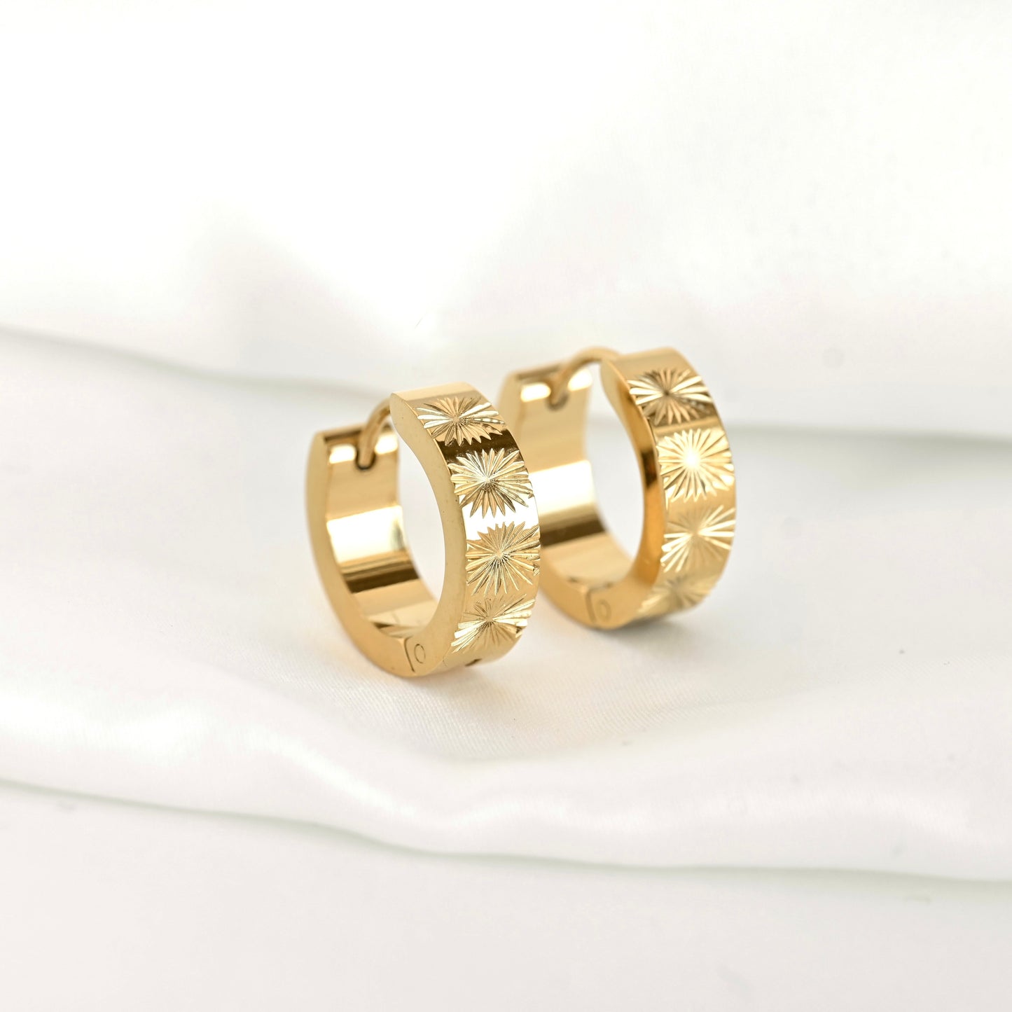 A pair of  small chunky gold hoop earrings with sunburst pattern on the front, displayed on white fabric.