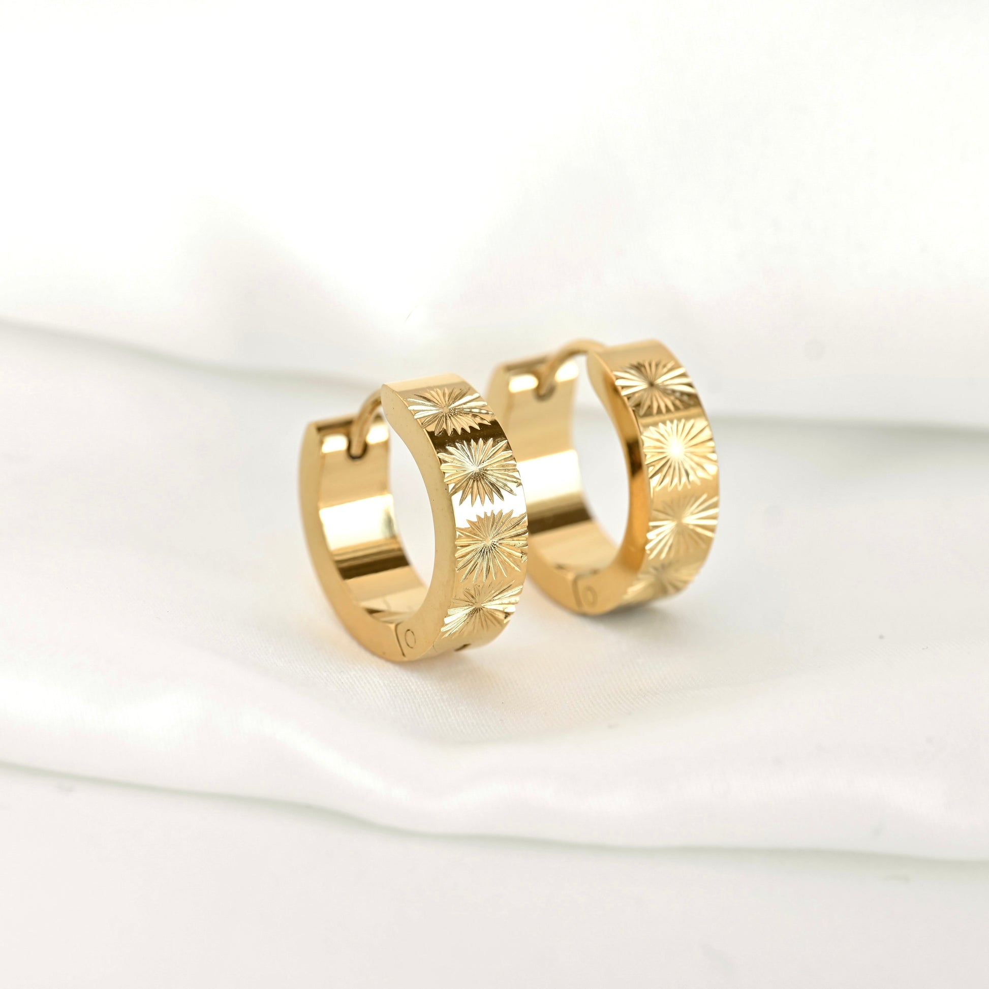A pair of  small chunky gold hoop earrings with sunburst pattern on the front, displayed on white fabric.