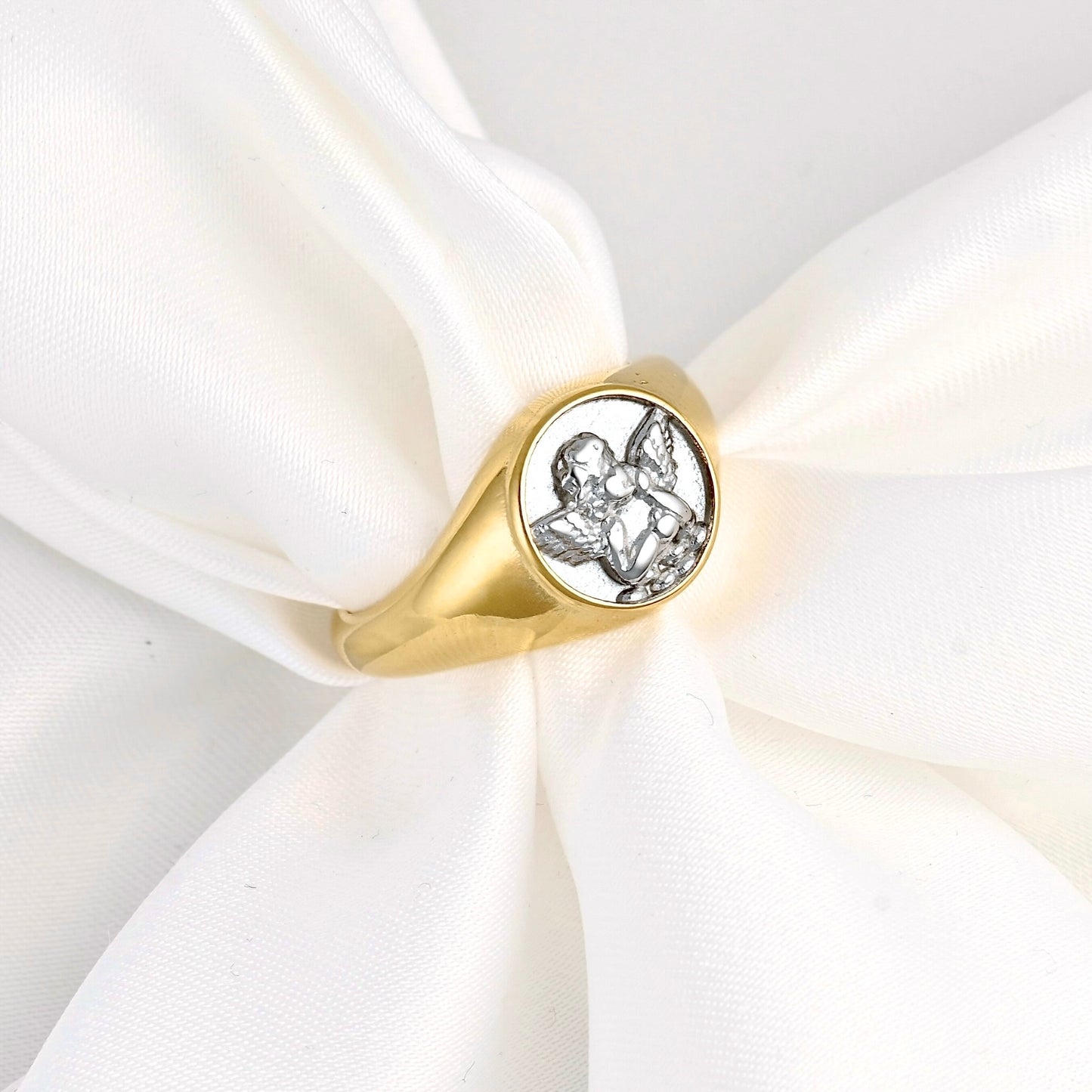 A gold signet ring with a silver-toned engraved angel design on a white satin ribbon.