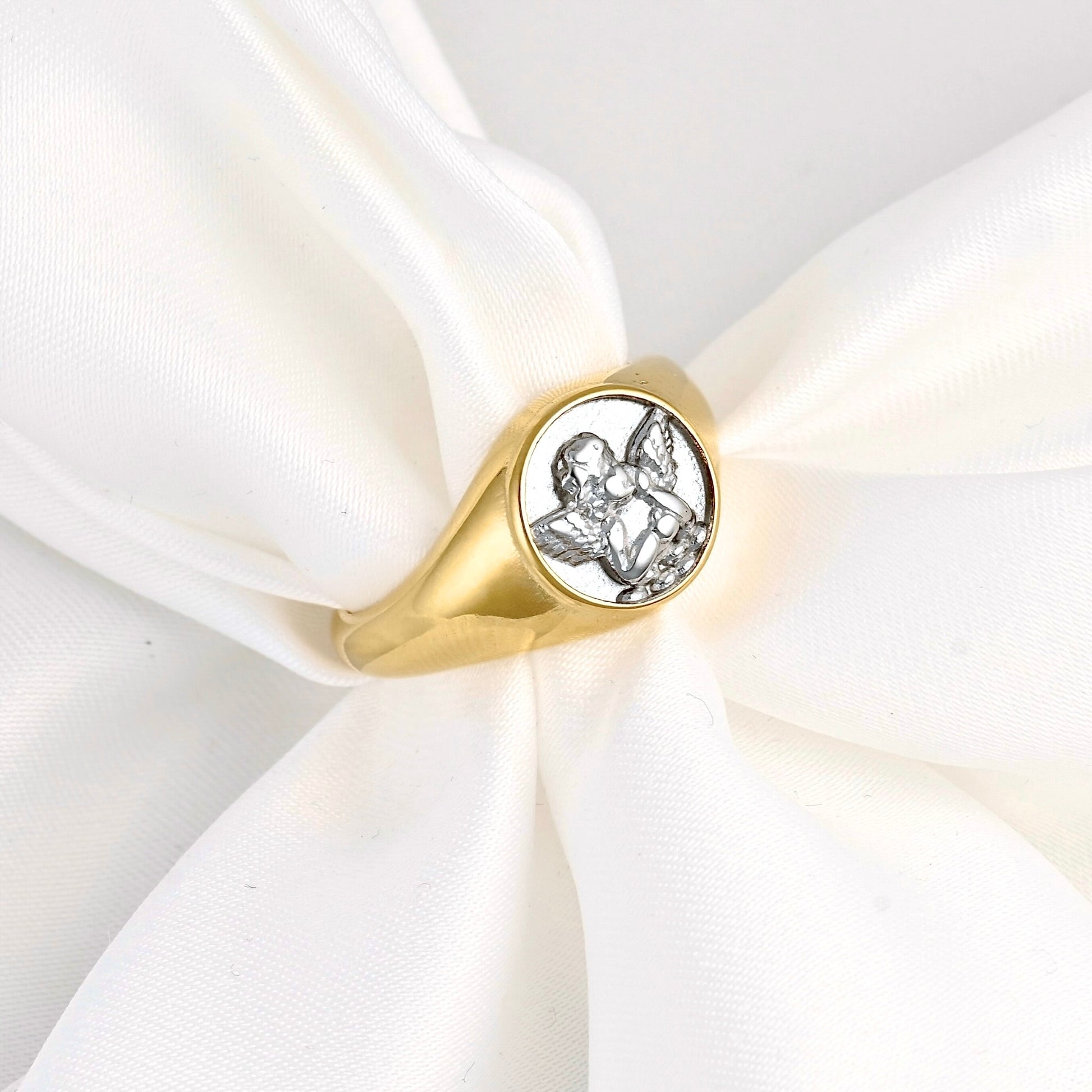 A gold signet ring with a silver-toned engraved angel design on a white satin ribbon.