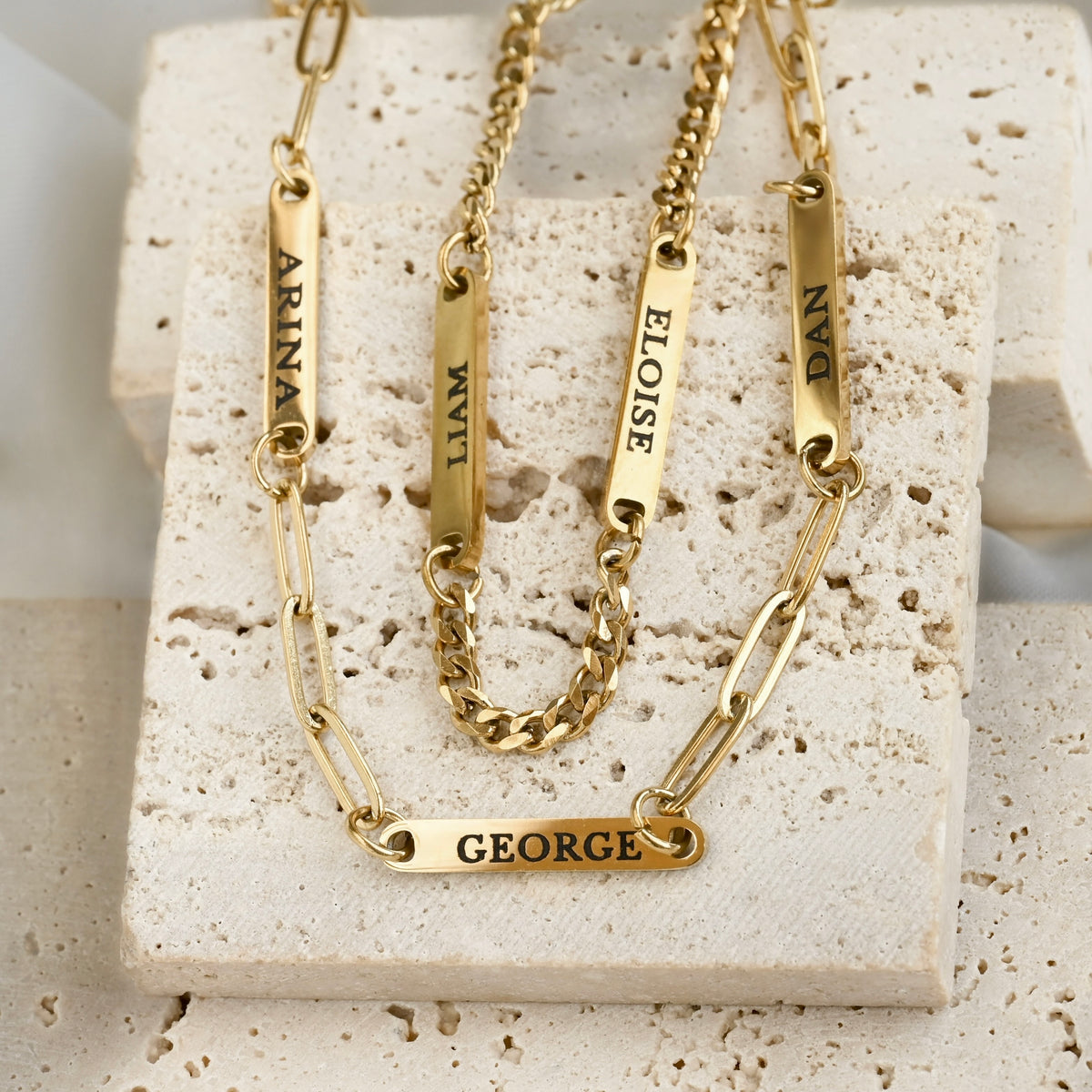 Two personalized gold nameplates on paperclip and curb chains, engraved with the names ARINA, WIL, ELOISE, DAN, and GEORGE, displayed on a textured beige stone block.