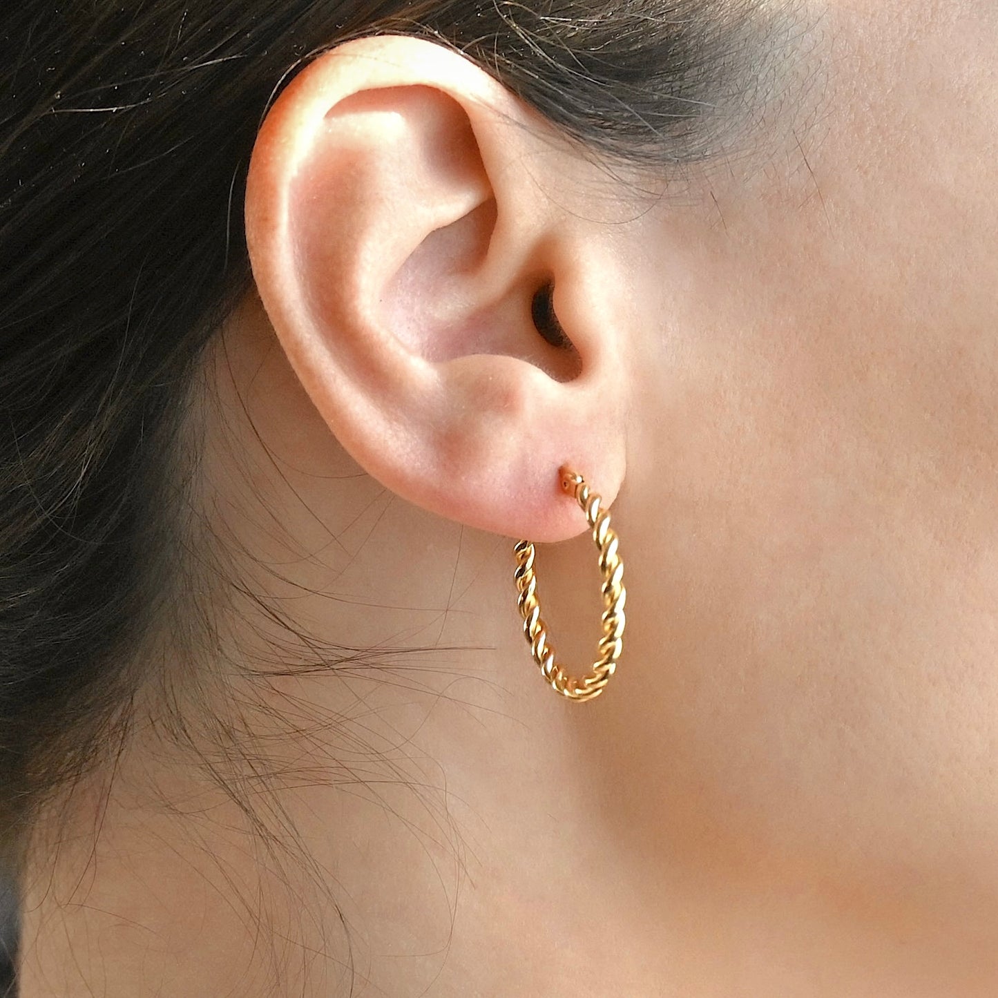 Close-up of an ear wearing a gold twisted hoops with blurred hair and skin in the background