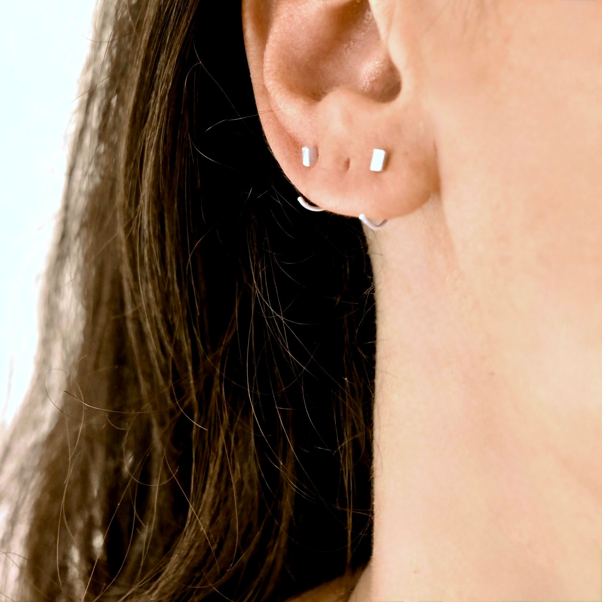 Close-up of a person's ear wearing two small silver open ear jacket hoop earrings, with dark brown hair and light skin visible.
