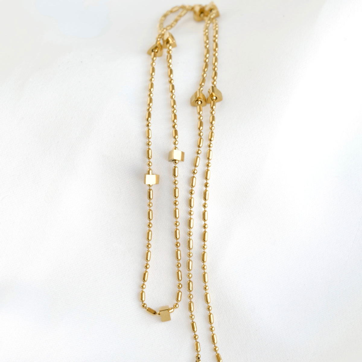 A delicate gold multi-strand necklace featuring small geometric cube and oval beads, laid out on a soft white fabric background.