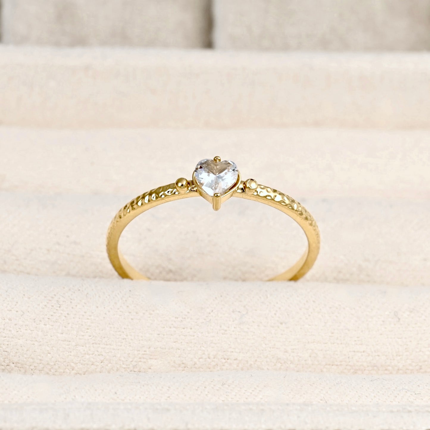 A gold ring with a heart-shaped clear gemstone in the center, displayed on a textured beige fabric ring stand.