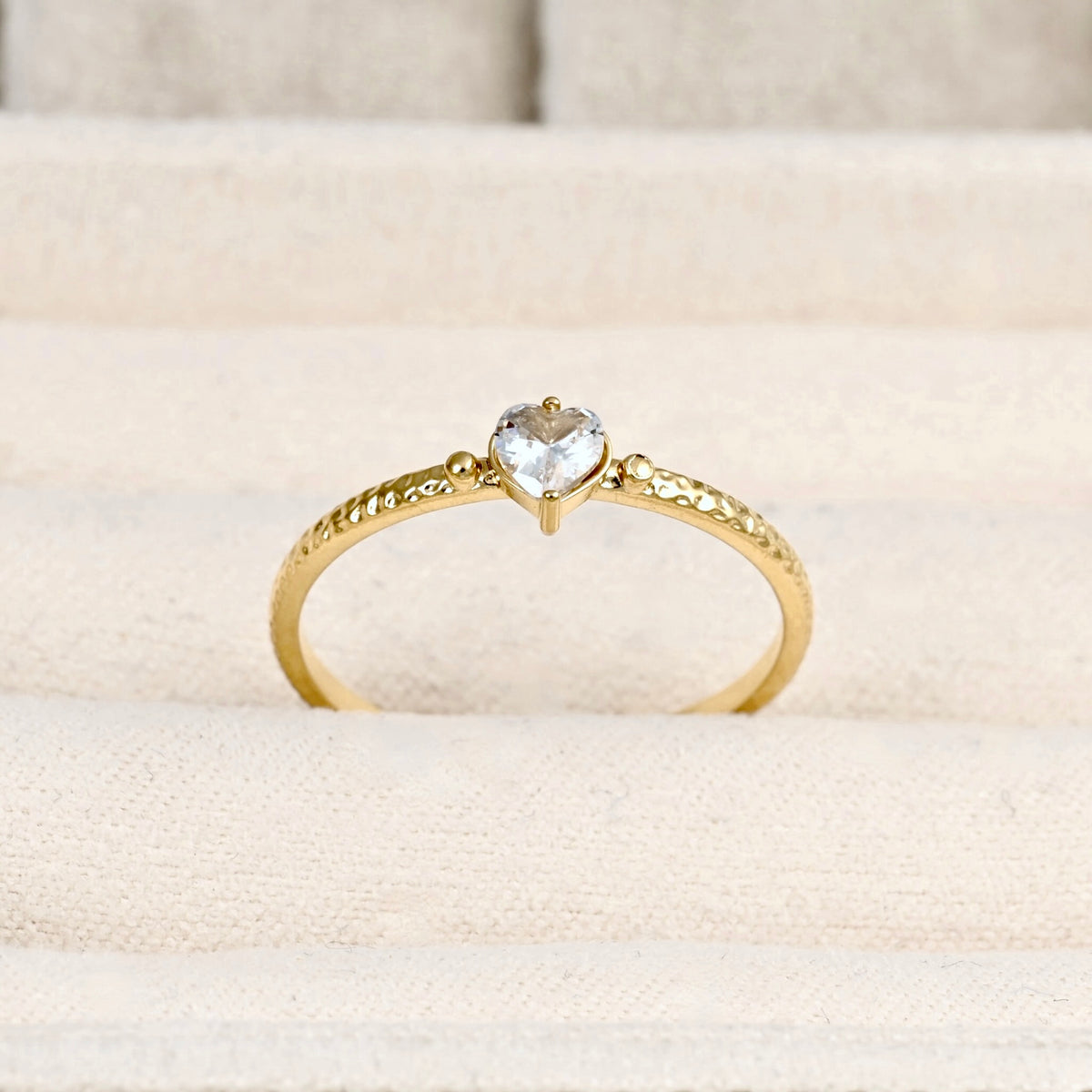 A gold ring with a heart-shaped clear gemstone in the center, displayed on a textured beige fabric ring stand.