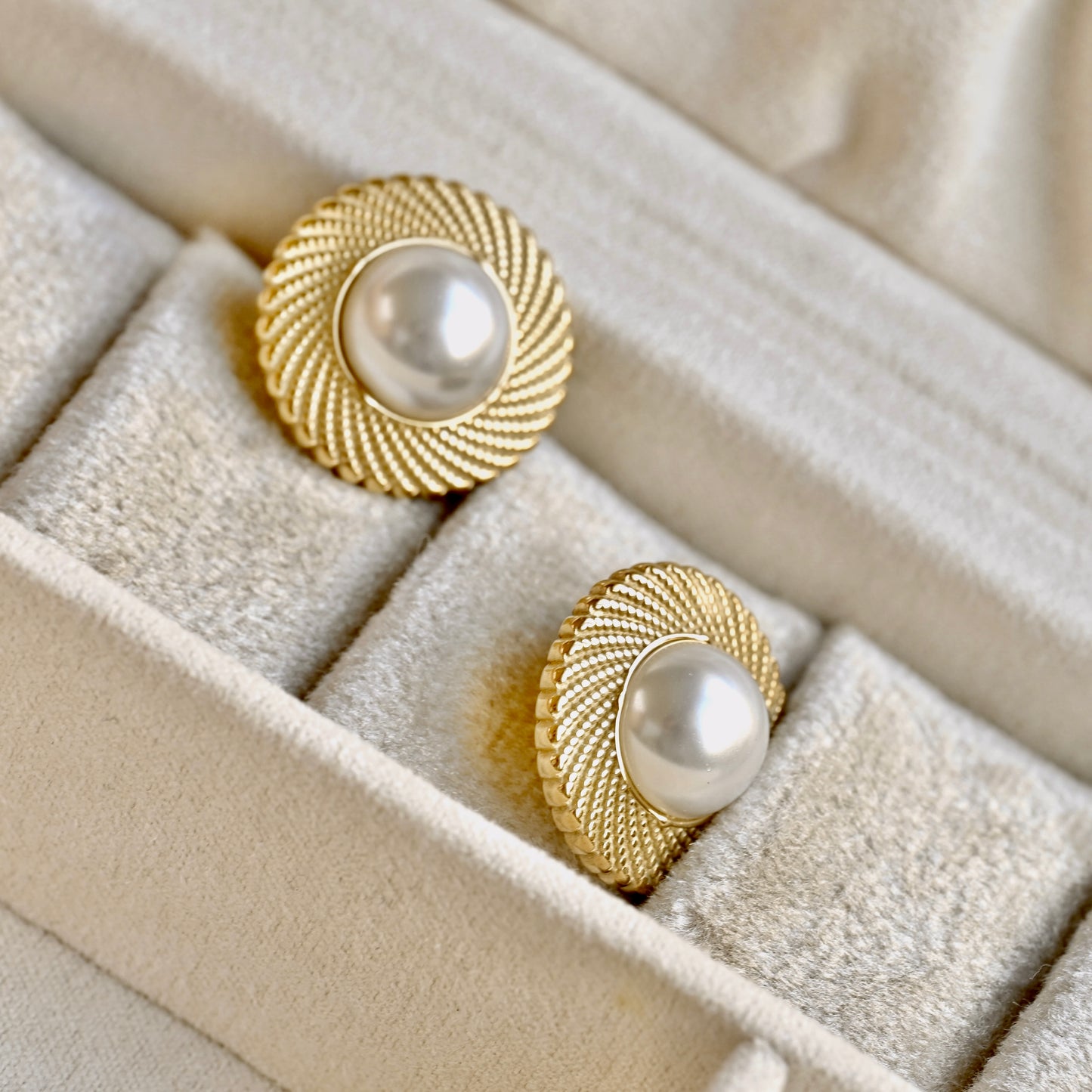 A pair of gold-toned round earrings with textured sunburst edges and white pearl centers, displayed in a beige velvet jewelry box.