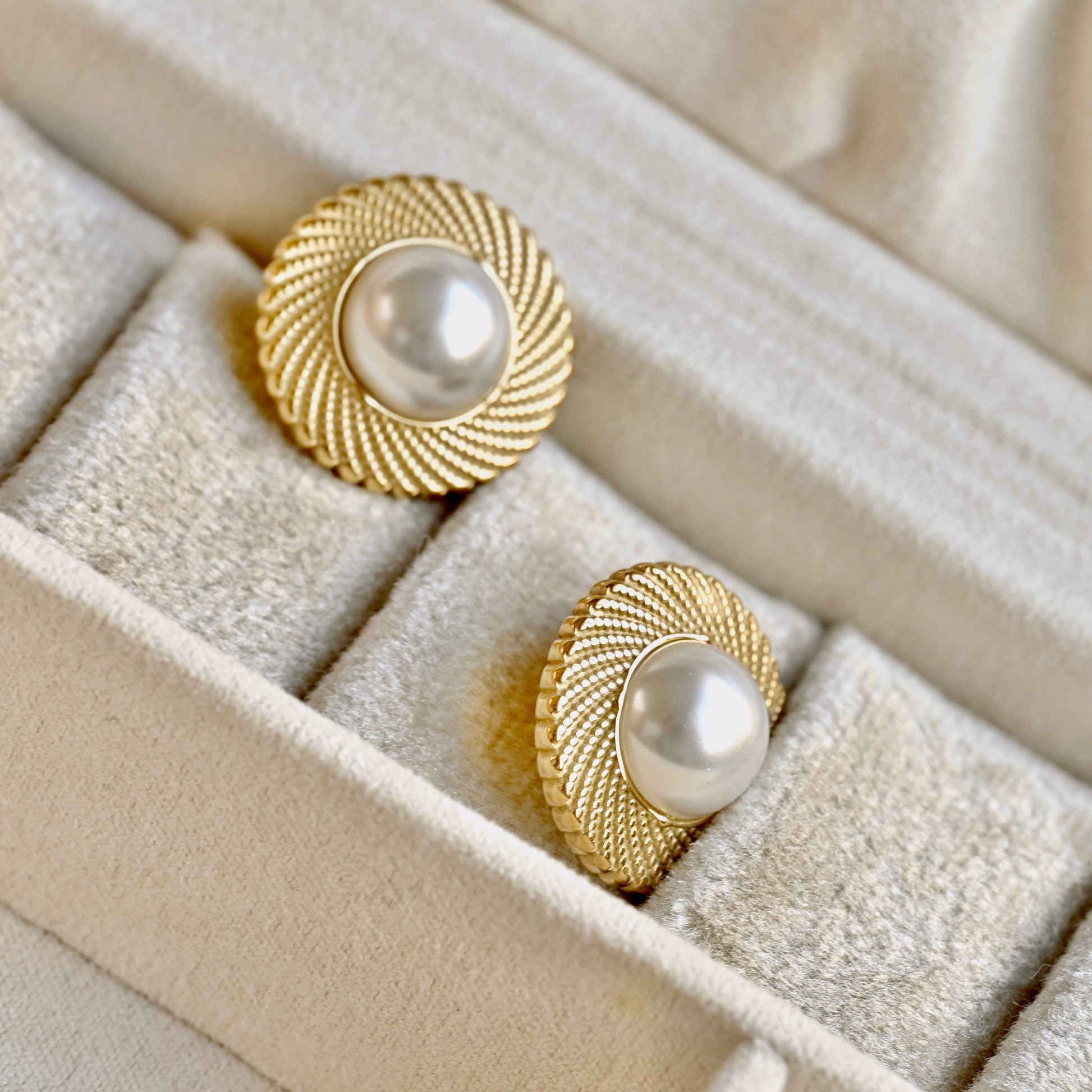 A pair of gold-toned round earrings with textured sunburst edges and white pearl centers, displayed in a beige velvet jewelry box.