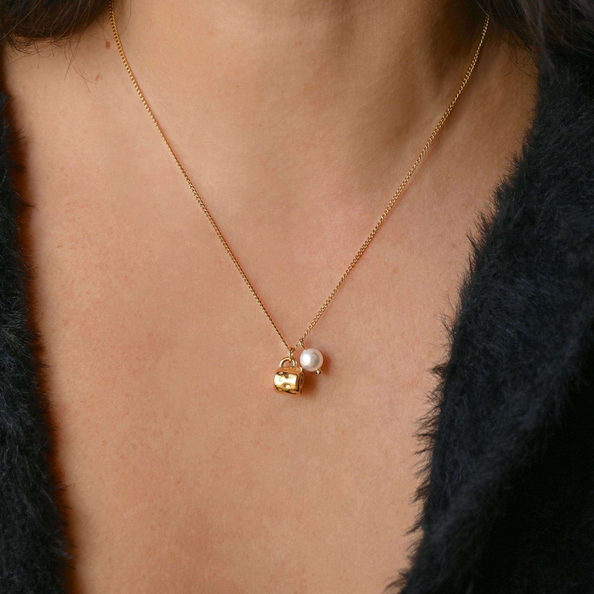 A close-up of a person wearing a delicate gold chain necklace with a small gold cup charm and a white pearl pendant, styled with a black fuzzy jacket.