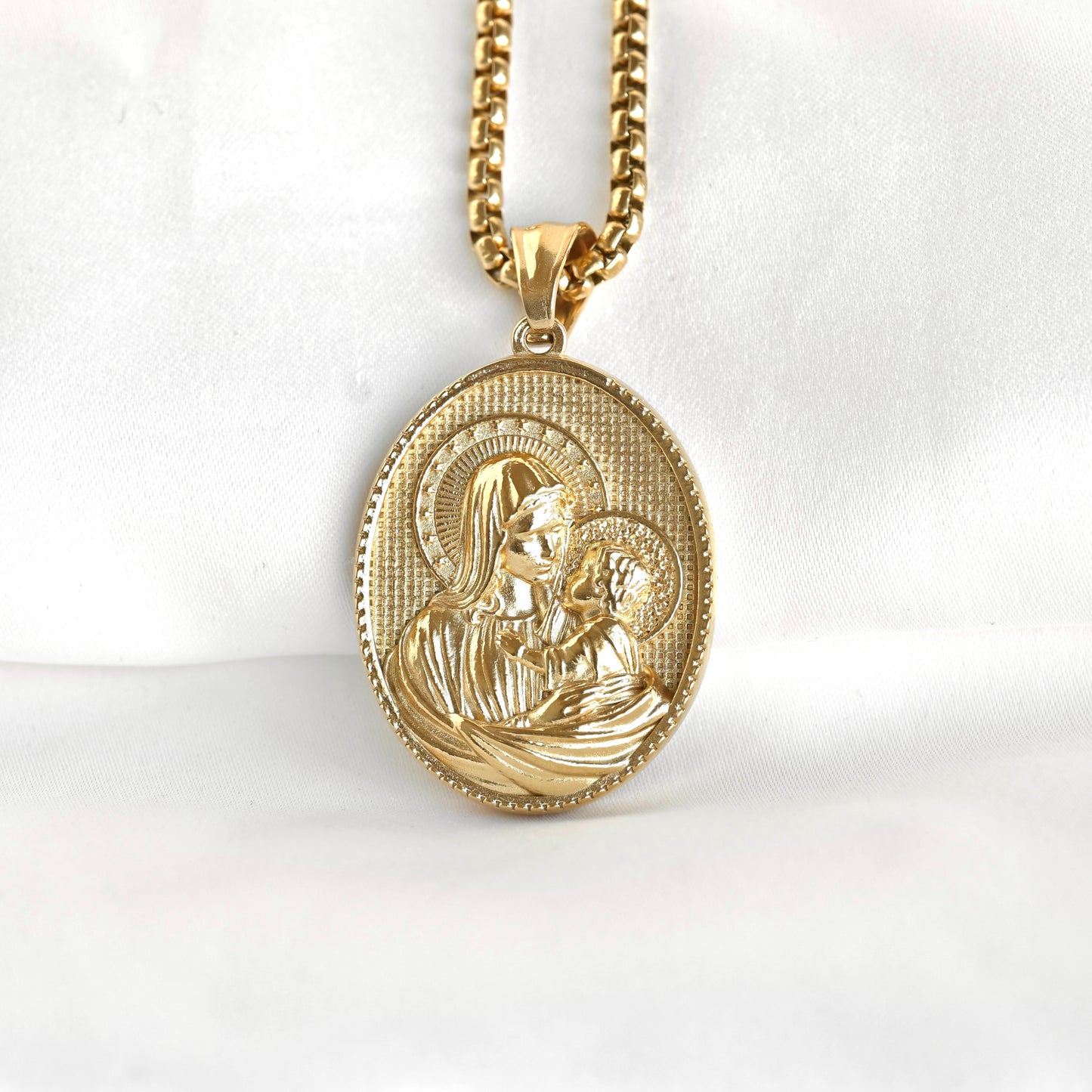 A gold oval religious pendant necklace featuring an embossed depiction of the Madonna and Child, suspended from a gold curb chain against a white fabric background.