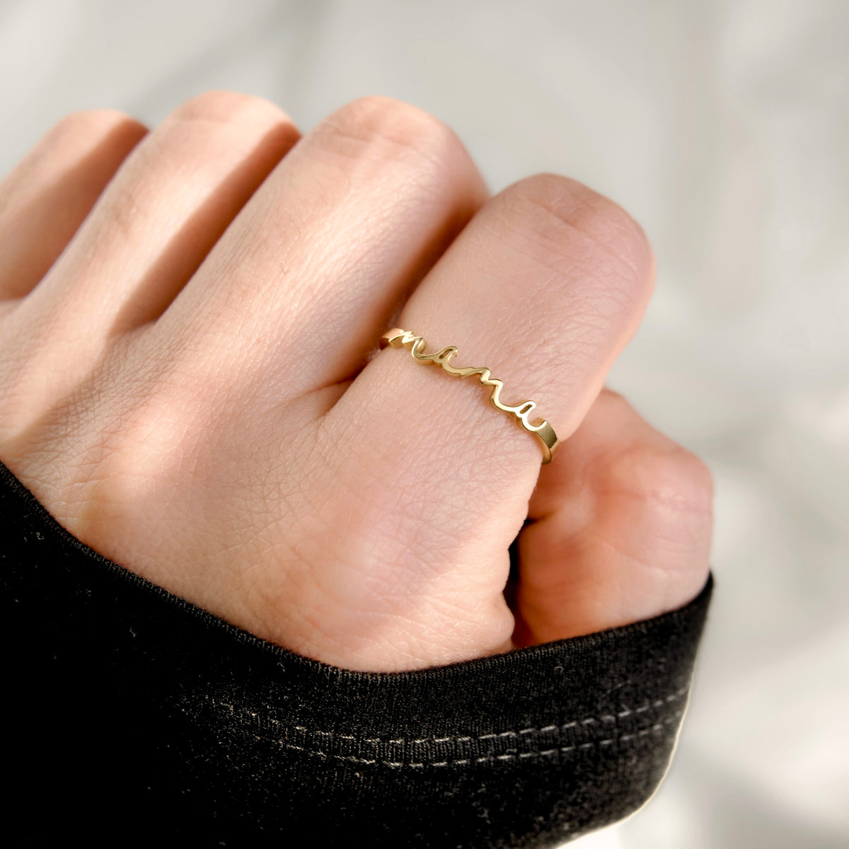 A close-up of a person's hand wearing a delicate gold ring with a wavy, script-like design on the ring finger.