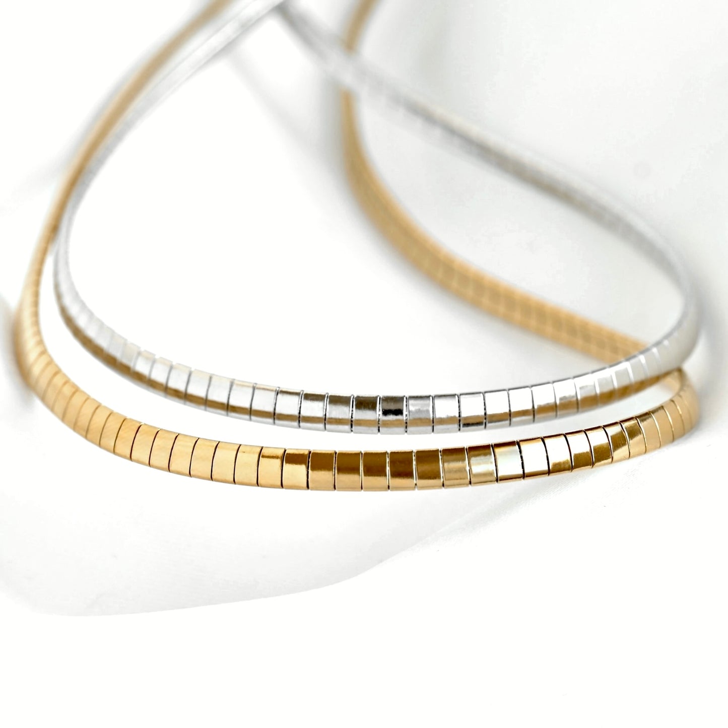 Two slim snake-bone style necklaces, one in polished yellow gold and one in polished silver, arranged in a layered loop on a clean white background.