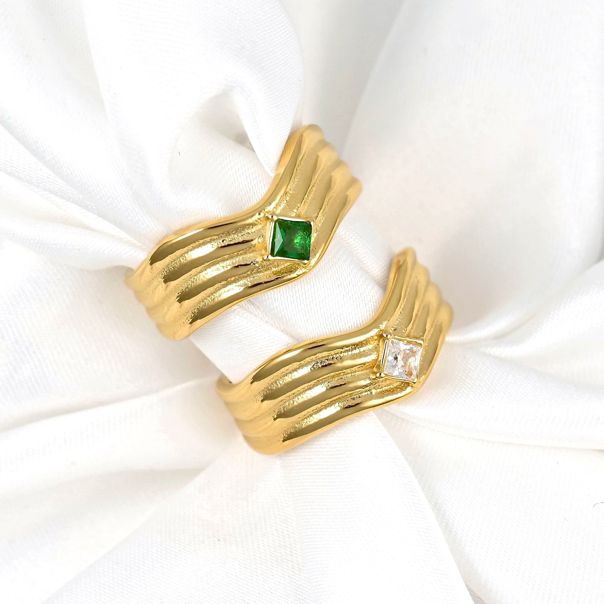 Two gold rings with textured, wavy bands displayed on a white fabric ring holder. One ring features a small square green gemstone, and the other features a small clear square gemstone.