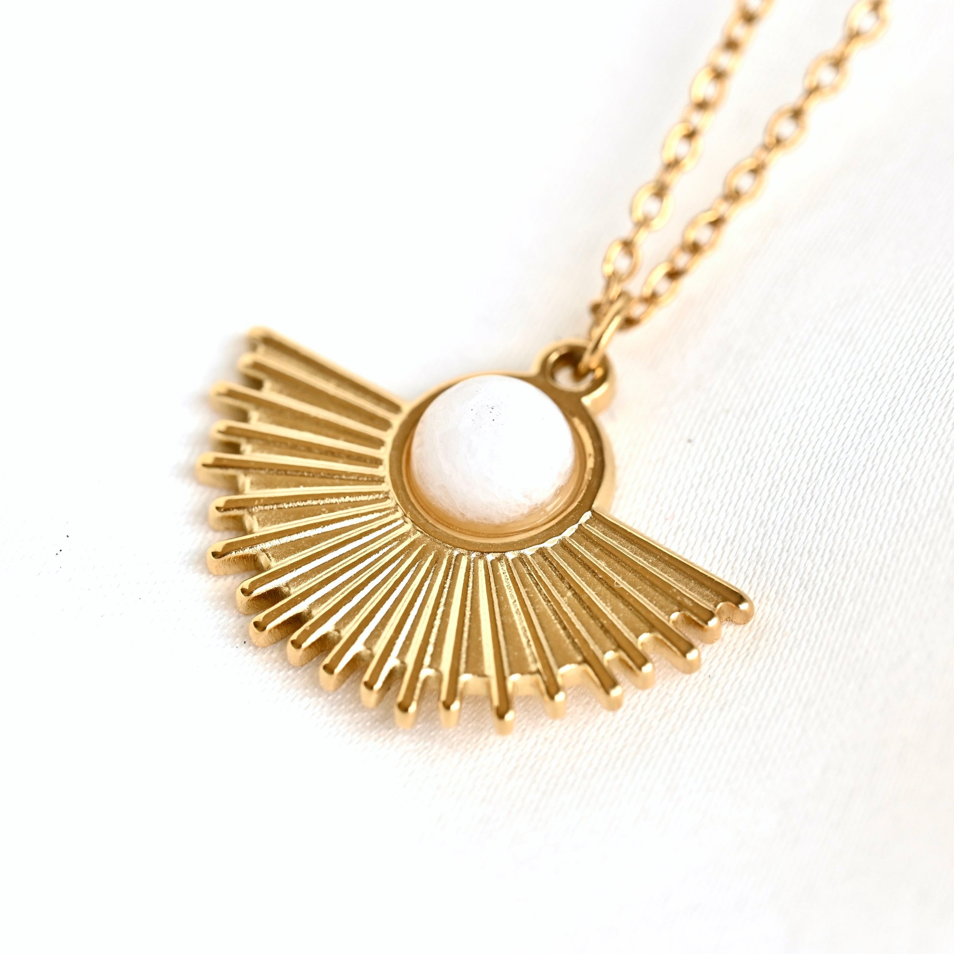 A gold pendant necklace featuring a sunburst fan design with a central white oval stone, shown on a white textured fabric background.
