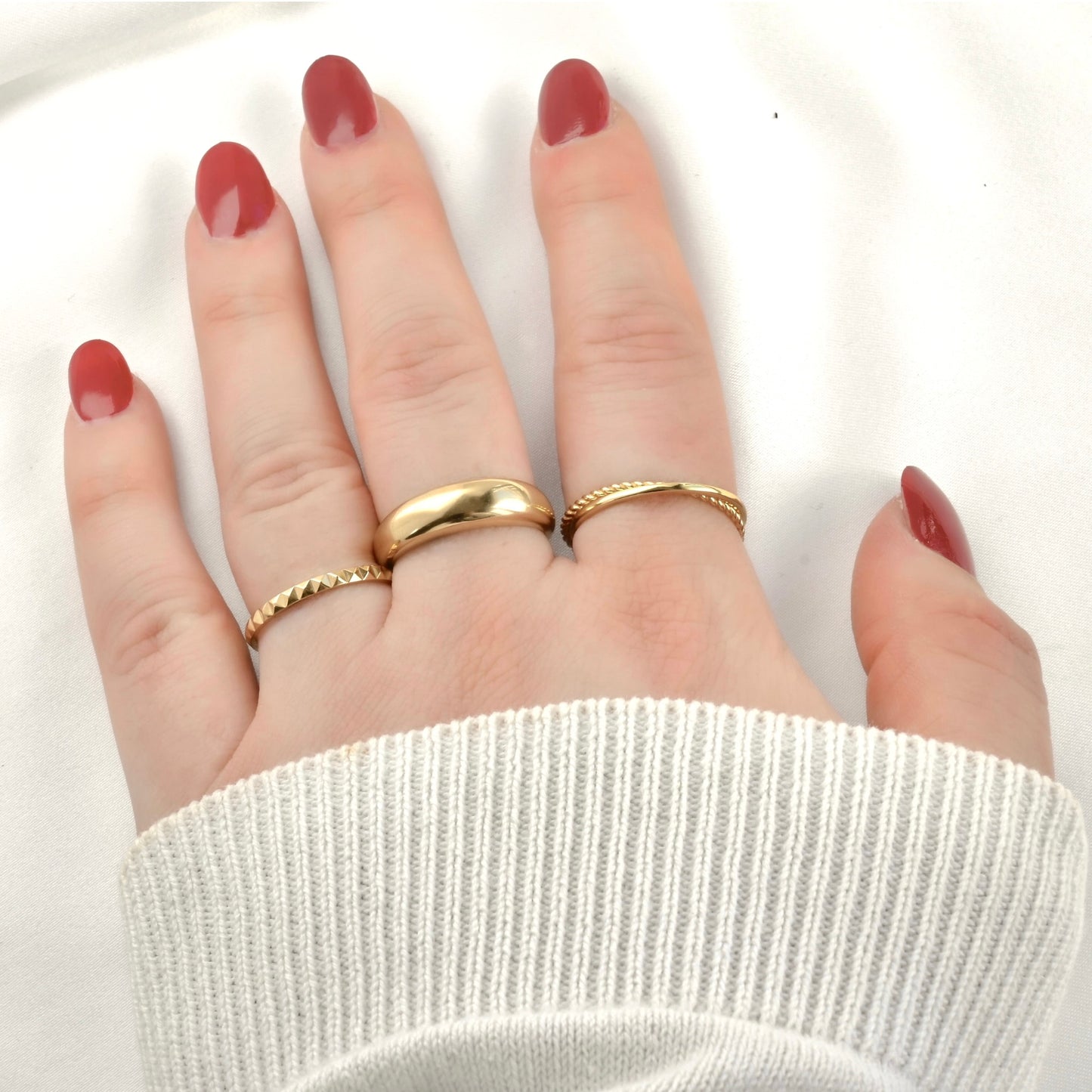 Hand wearing a stack of three gold rings with red nail polish on a white background