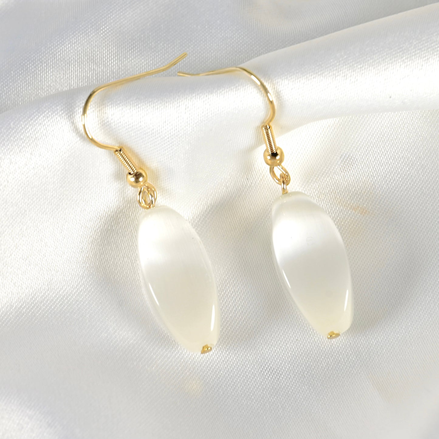 A pair of gold-tone drop earrings with smooth, teardrop-shaped cat eye's stone pendants, displayed on a white satin fabric background.