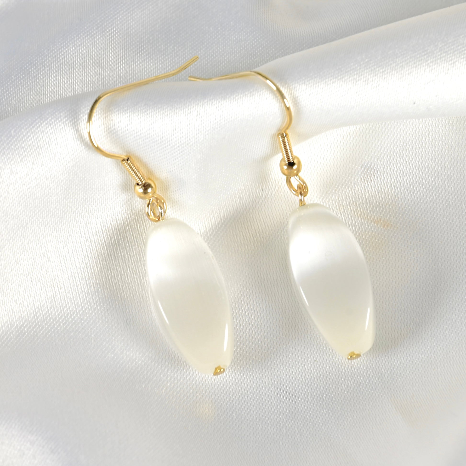 A pair of gold-tone drop earrings with smooth, teardrop-shaped cat eye's stone pendants, displayed on a white satin fabric background.