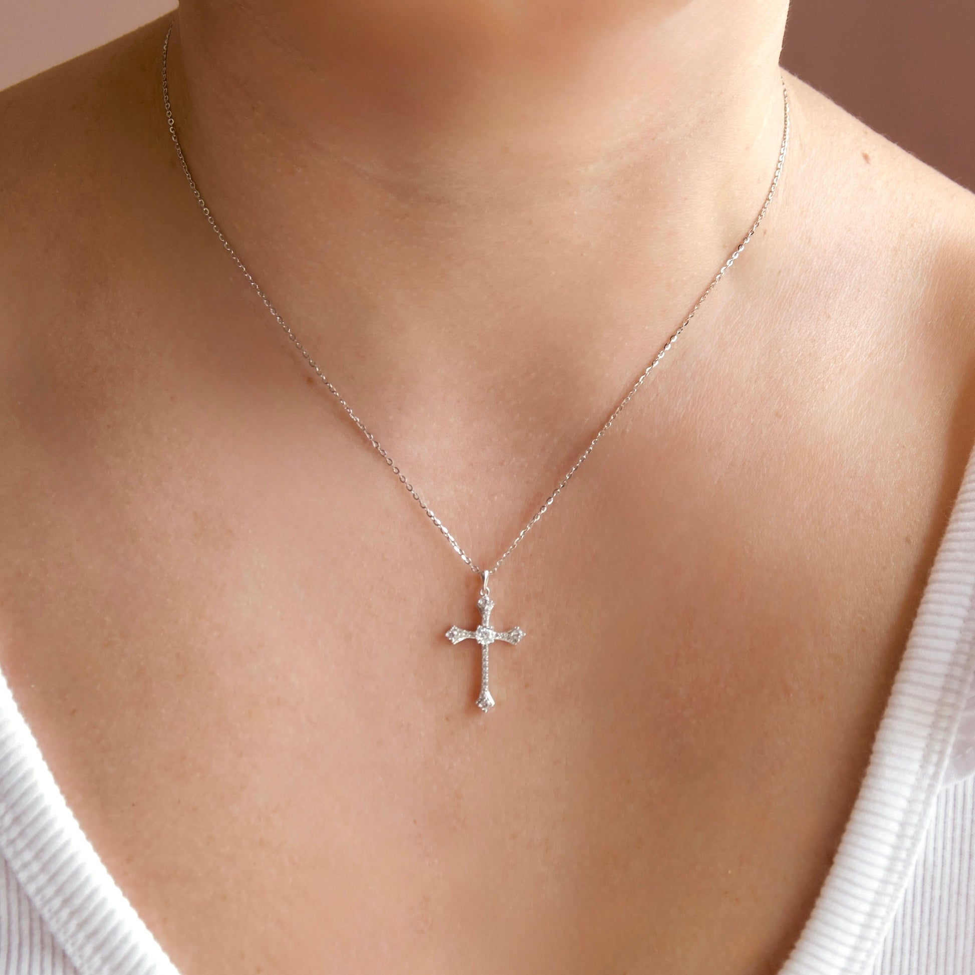 A delicate silver cross pendant necklace worn on a model's neck, featuring small clear crystals set along the arms of the cross, paired with a white ribbed top.