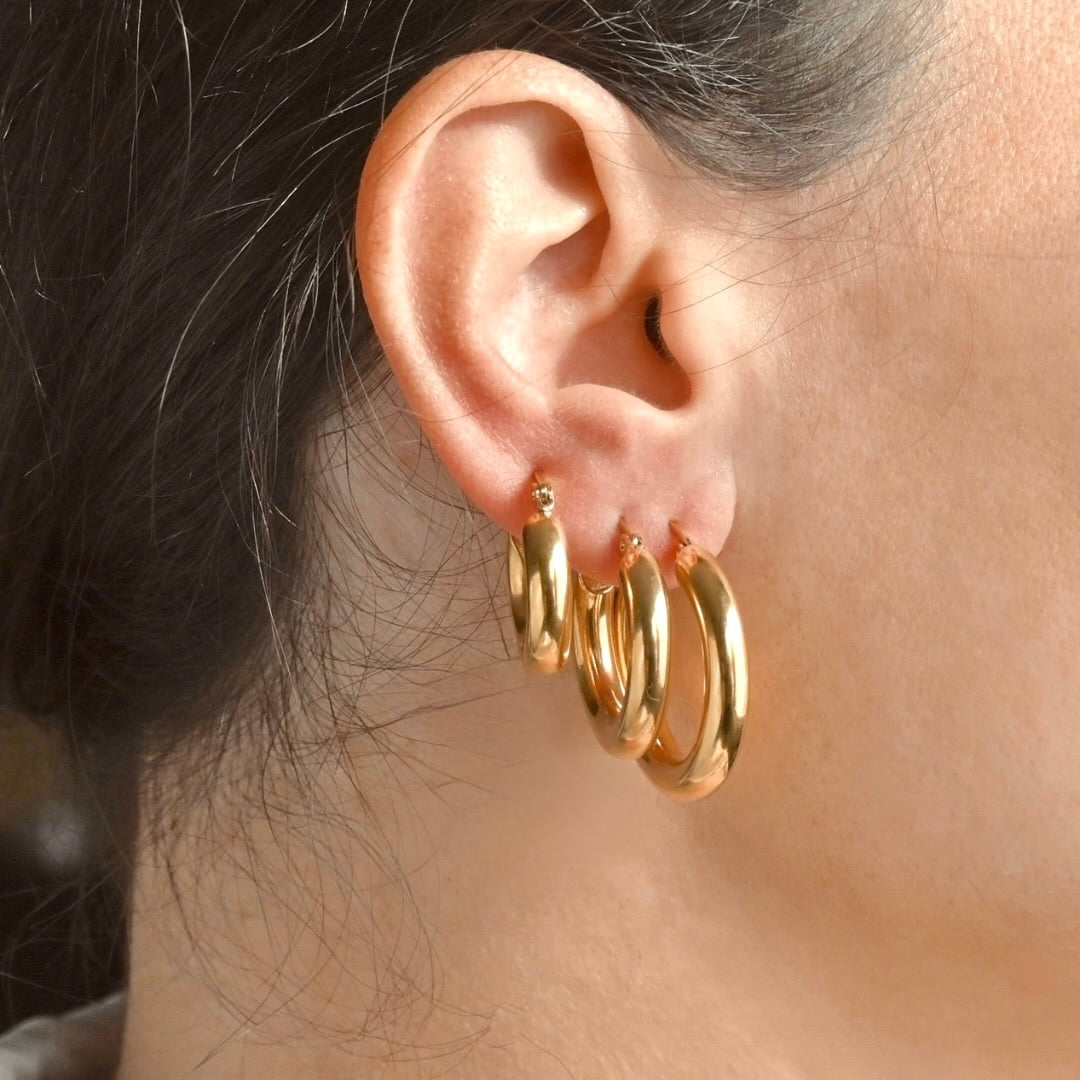 Close-up of an ear wearing three thick gold hoop earrings in three sizes: 20mm, 25mm and 30mm, with a blurred background