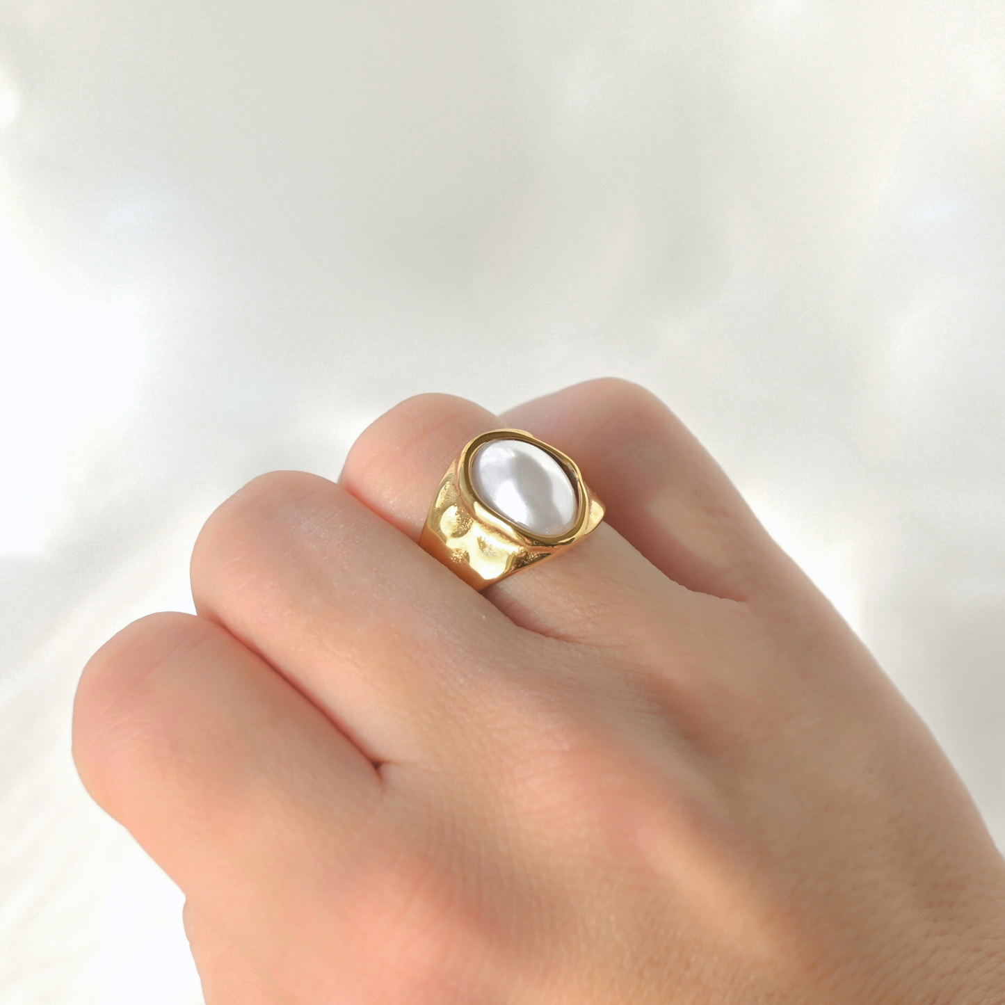 A gold signet-style ring with a large white pearl-like oval center, worn on a person's finger against a soft white fabric background.