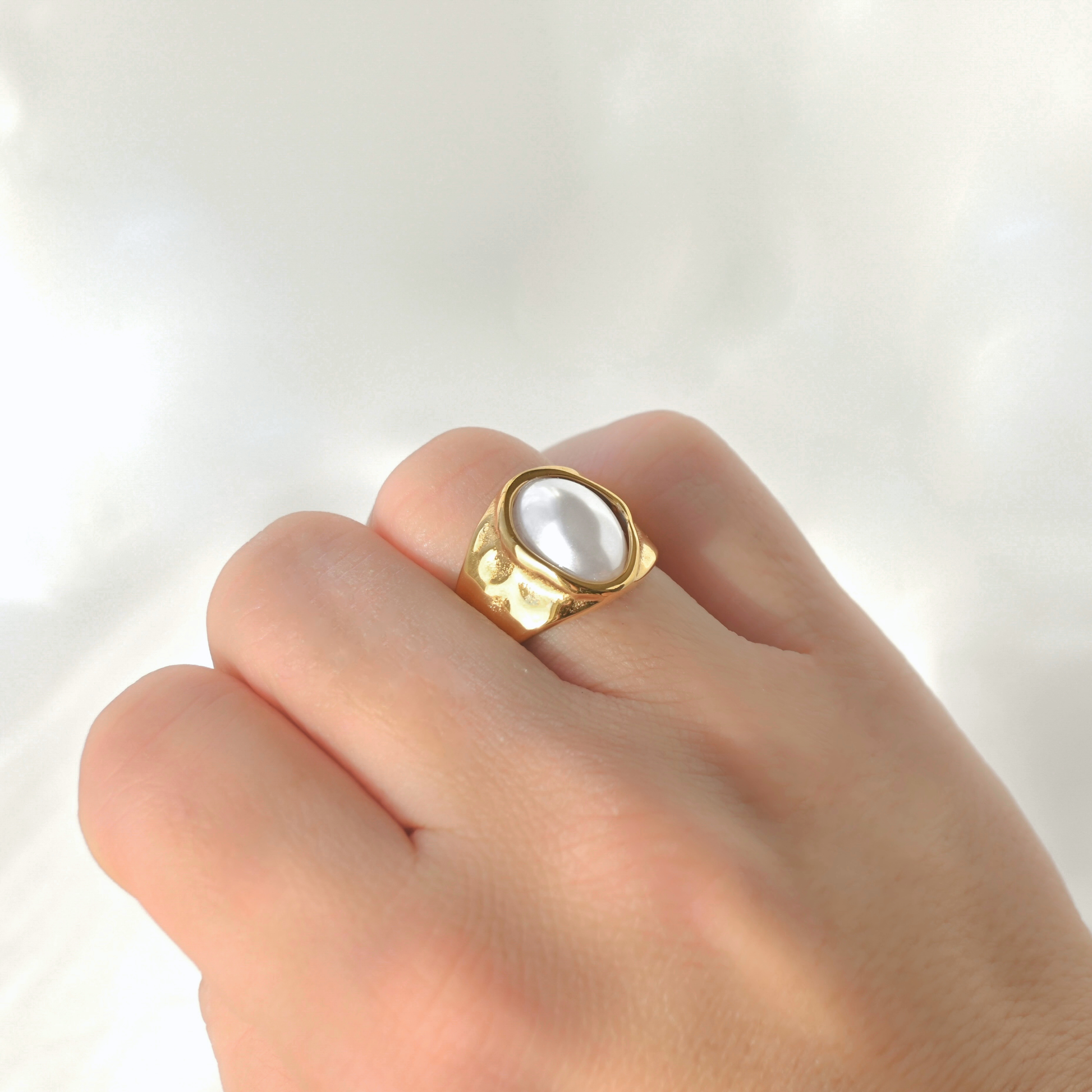 A gold signet-style ring with a large white pearl-like oval center, worn on a person's finger against a soft white fabric background.
