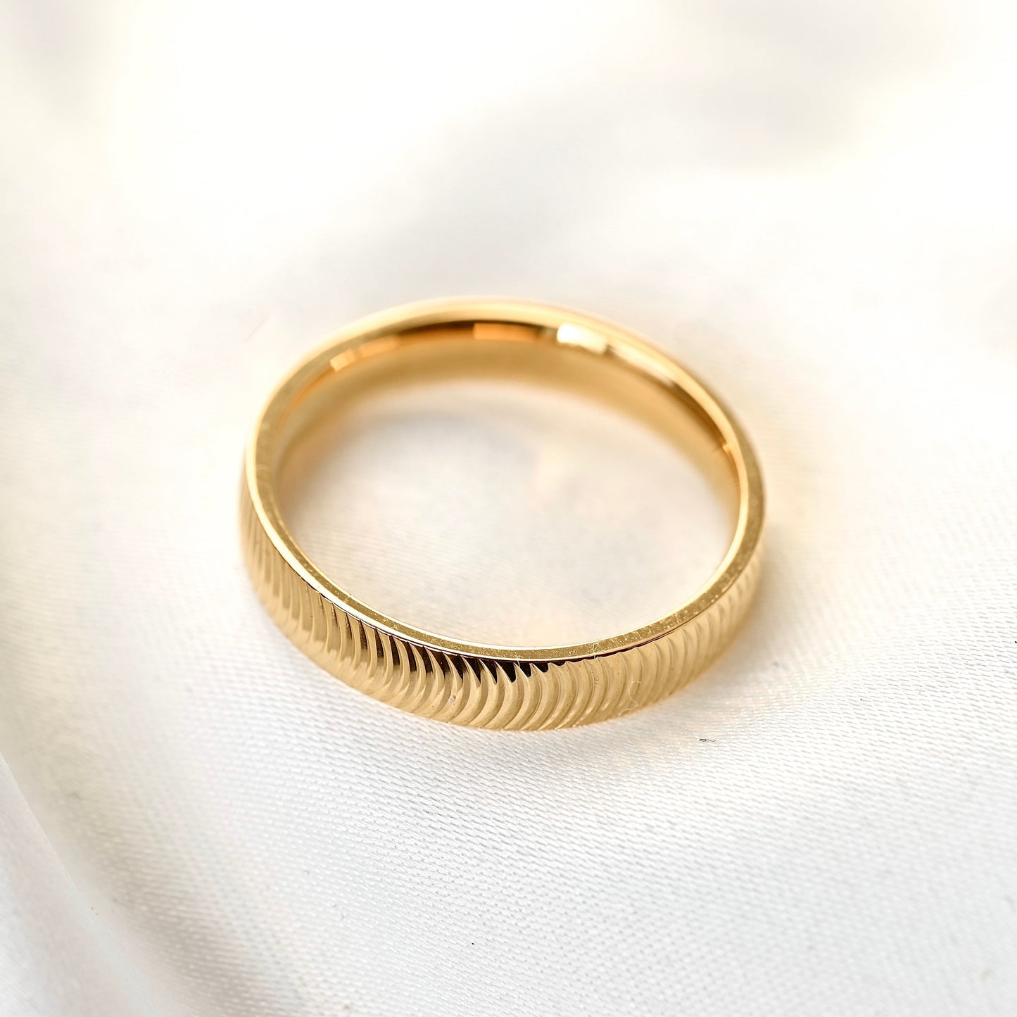 A gold band ring with a textured, ribbed exterior and a polished inner band, resting on a soft white fabric background.