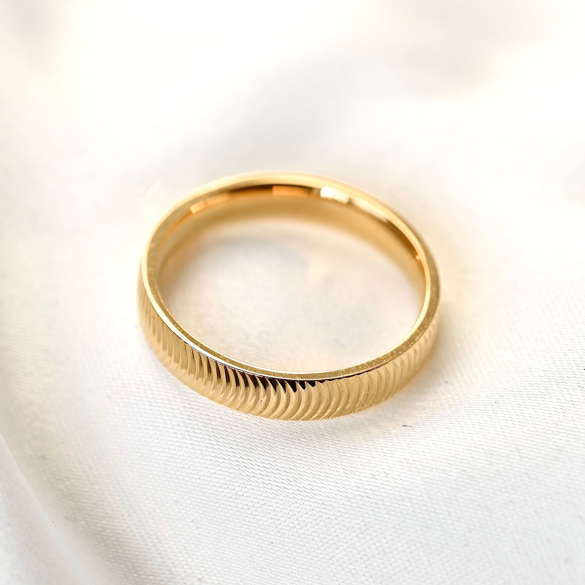 A gold band ring with a textured, ribbed exterior and a polished inner band, resting on a soft white fabric background.