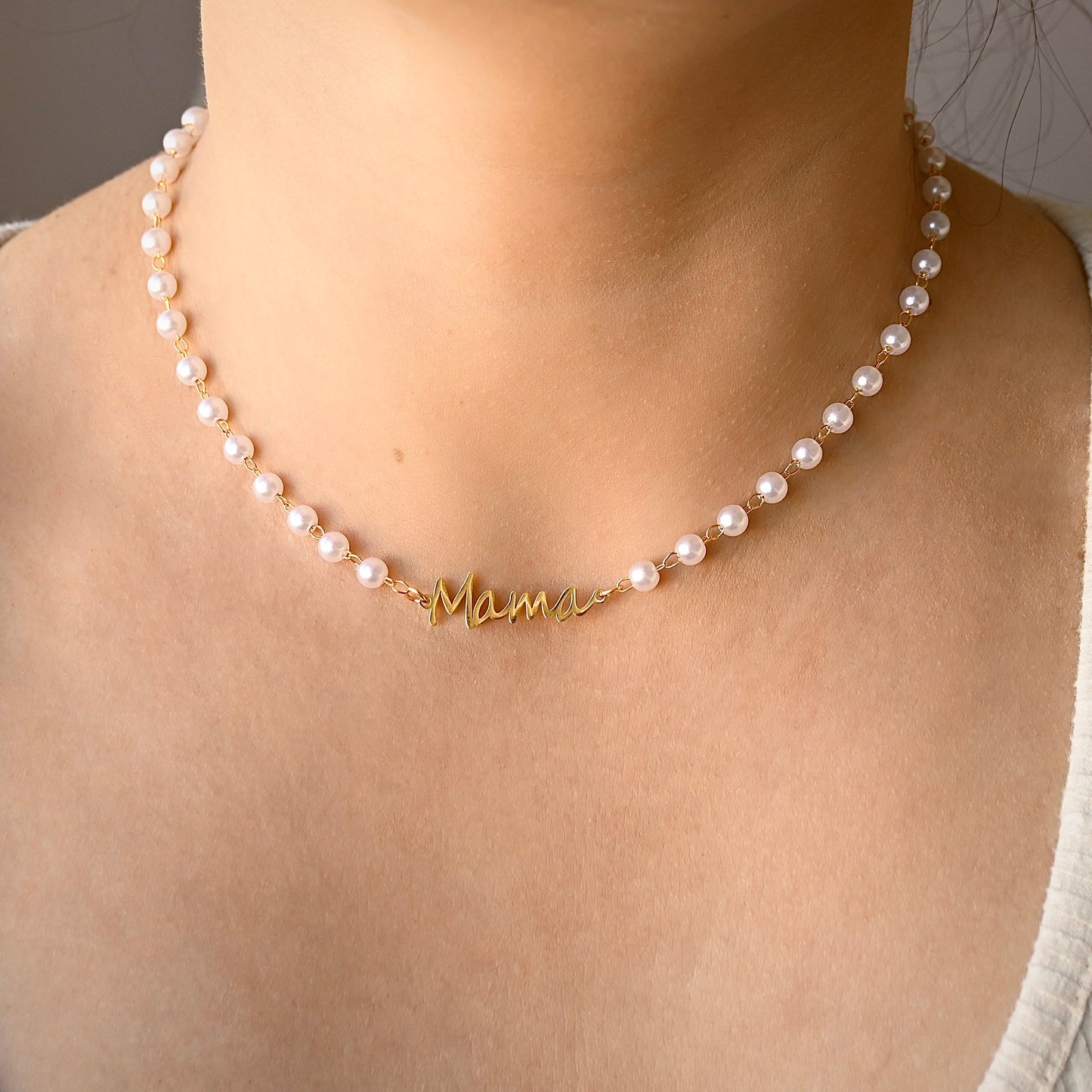 A close-up of a person's neck and upper chest wearing a delicate necklace made of small white pearls with a gold script nameplate reading 'Mami'.
