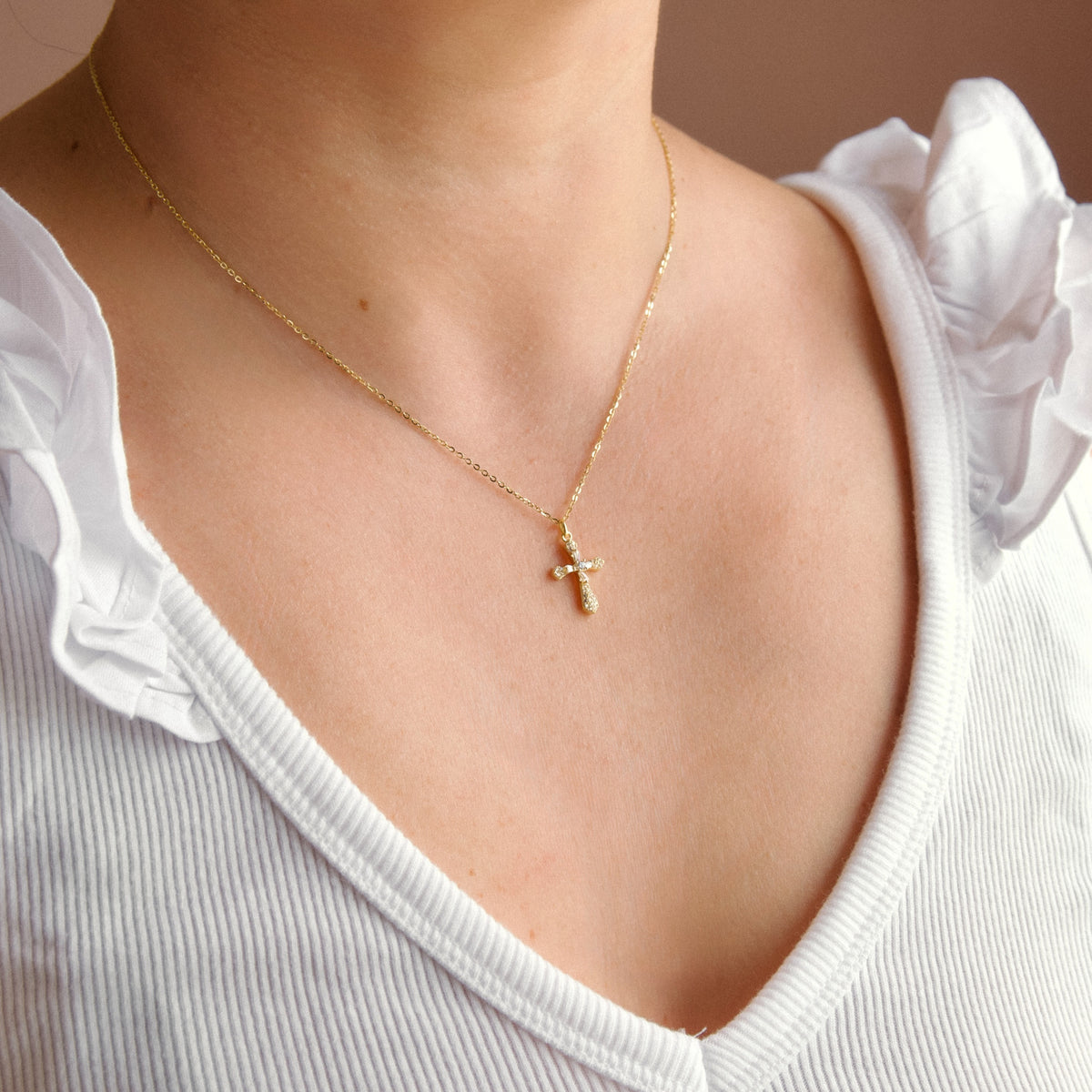 A close-up of a person wearing a delicate gold cross pendant necklace on a fine chain, styled with a white ruffled top.