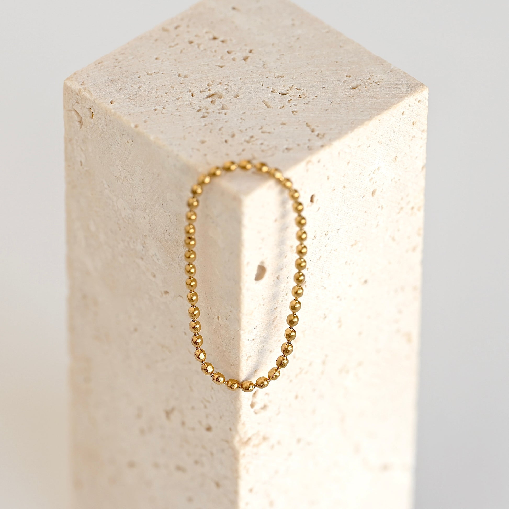 A delicate gold beaded necklace draped over a textured beige stone against a clean white background.