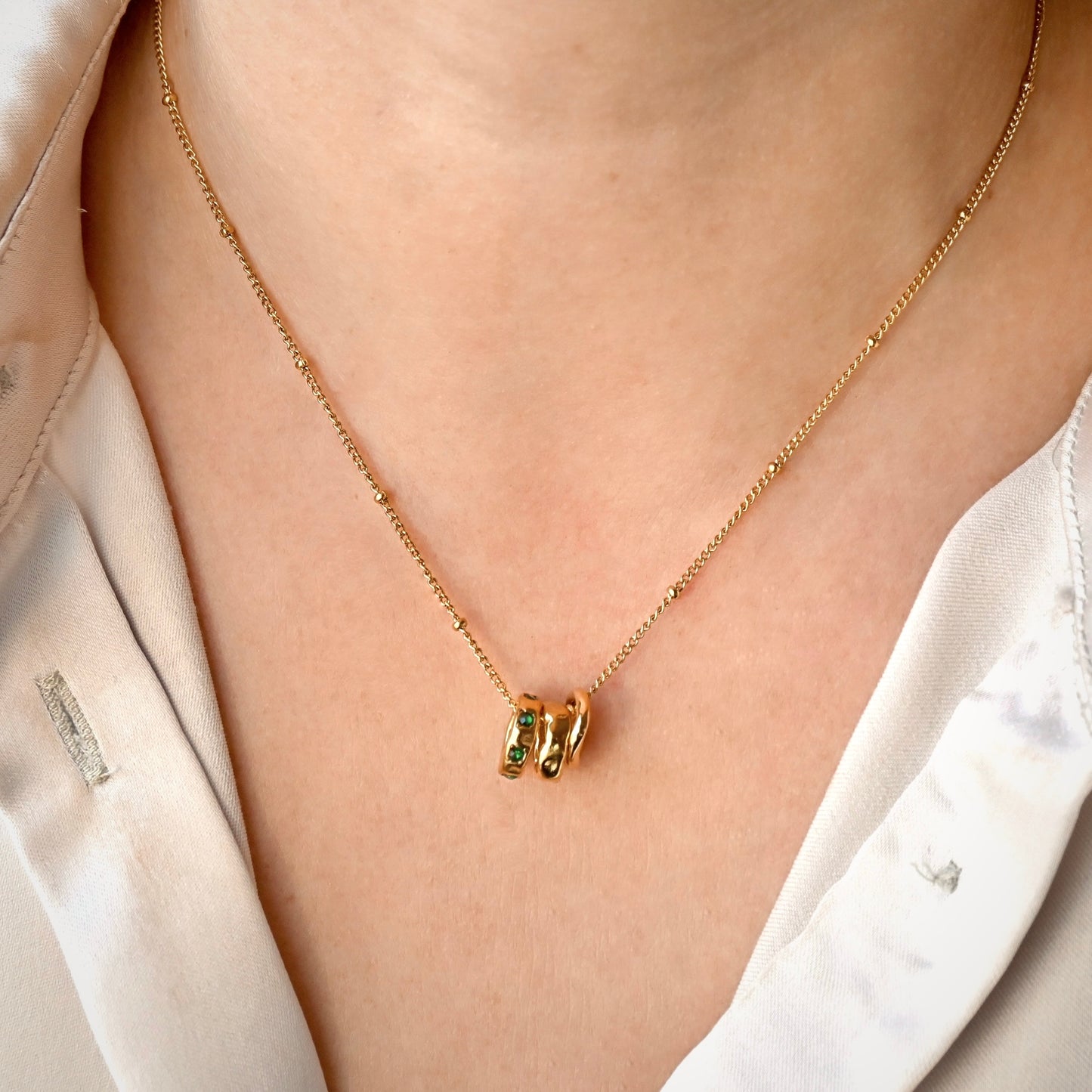 A close-up of a person wearing a delicate gold satellite chain necklace with three small textured rings, one of them accented with green gemstones, styled with a white collared shirt.