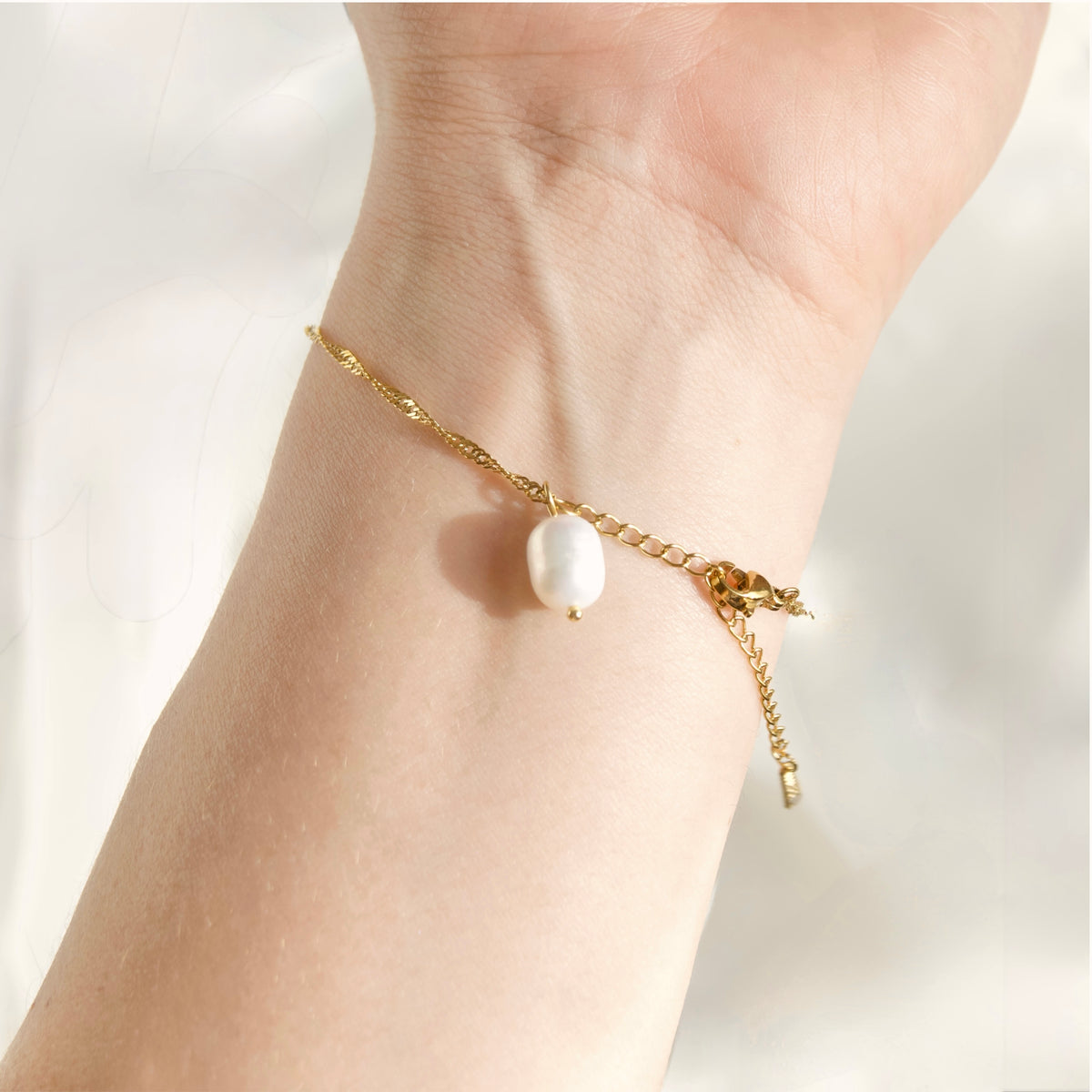 A delicate gold chain bracelet with a single white freshwater pearl charm, shown worn on a person's wrist against a bright, soft-focus background.