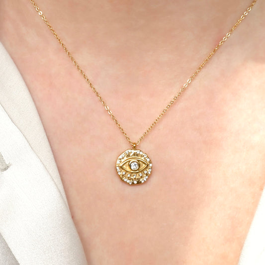 Gold necklace with an eye-shaped coin pendant adorned with clear stones on a person wearing a white shirt.
