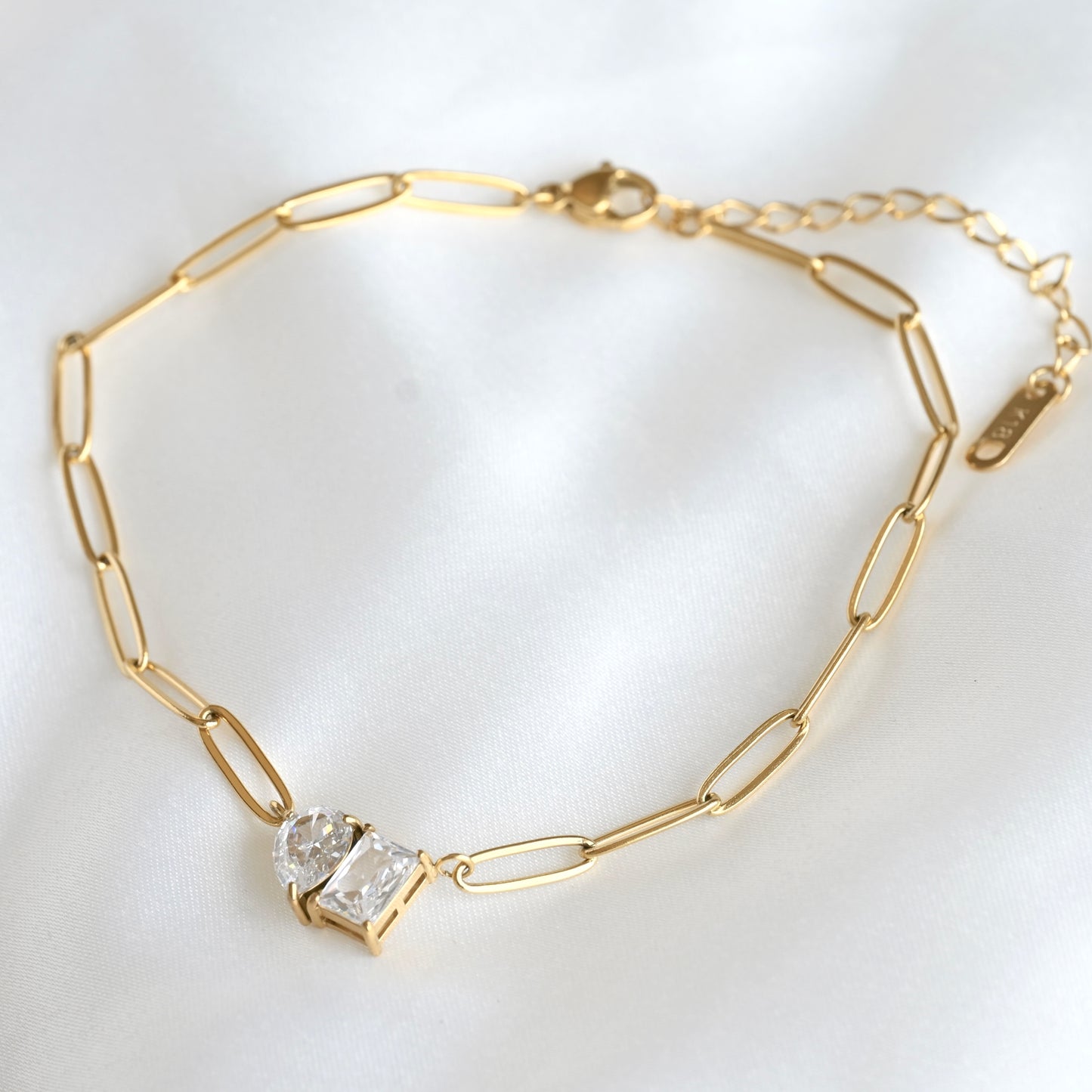 A gold paperclip chain necklace with a prong-set clear gemstone pendant, displayed on a soft white fabric background.