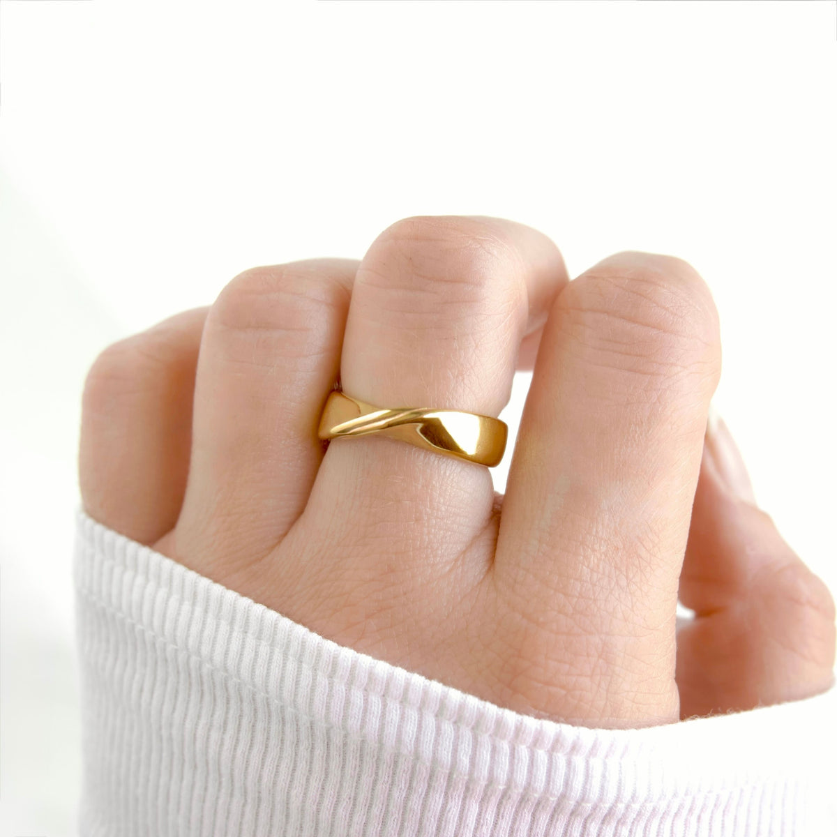 A close-up of a hand wearing a polished gold ring with a twisted, fluid design, styled with a white ribbed knit sleeve against a bright white background.